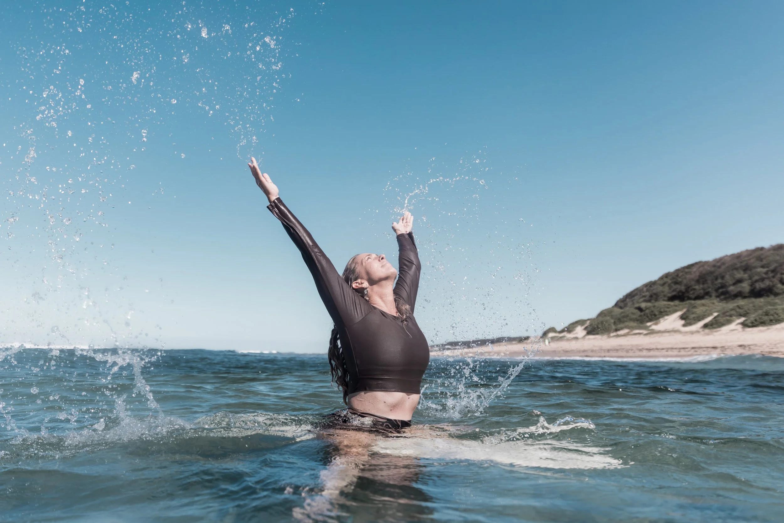 A woman in a black wetsuit standing in the ocean with arms raised, splashing water above her head, at the beach on a sunny day with cliffs in the background.
