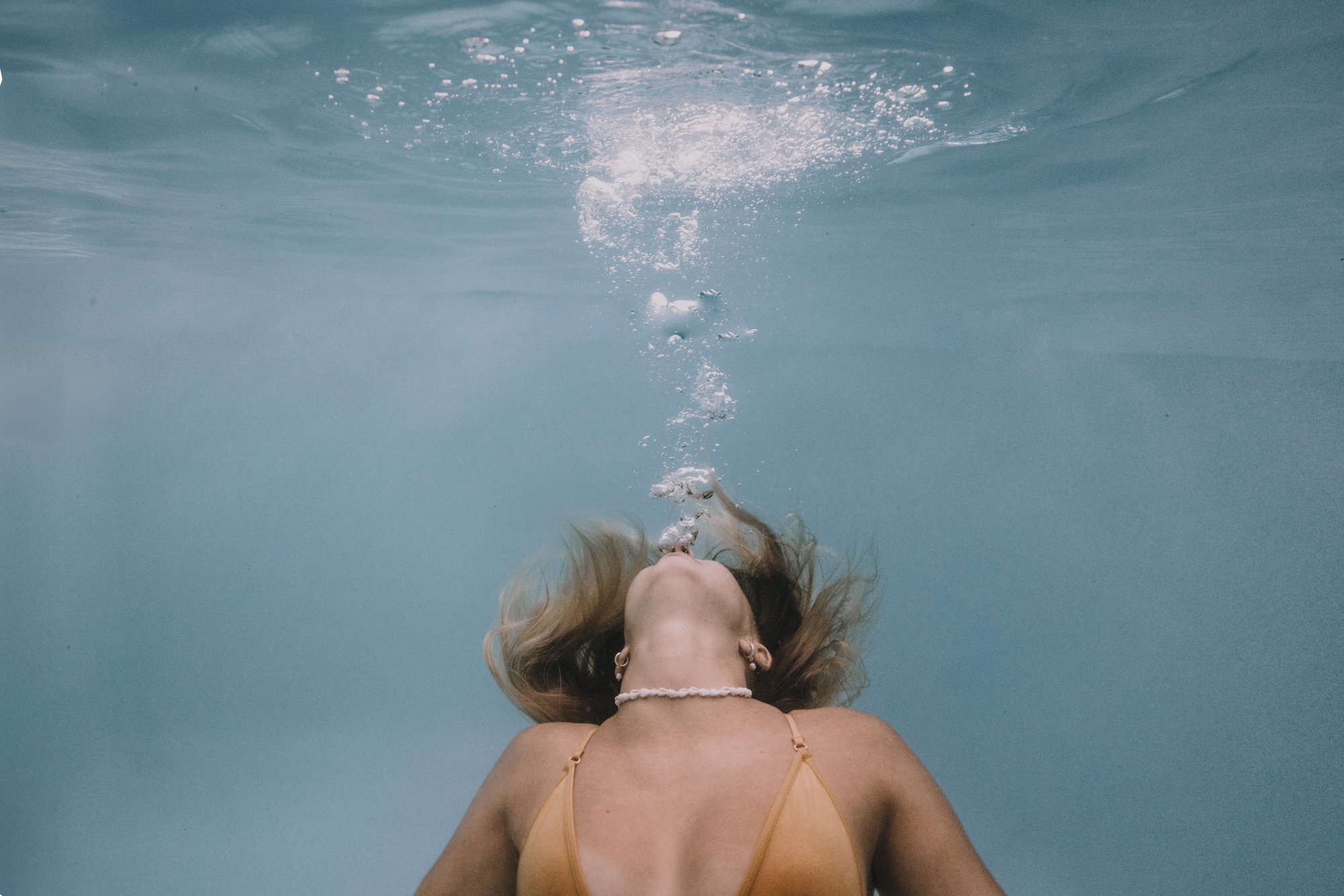 Person underwater blowing bubbles, wearing a swimsuit and necklace.