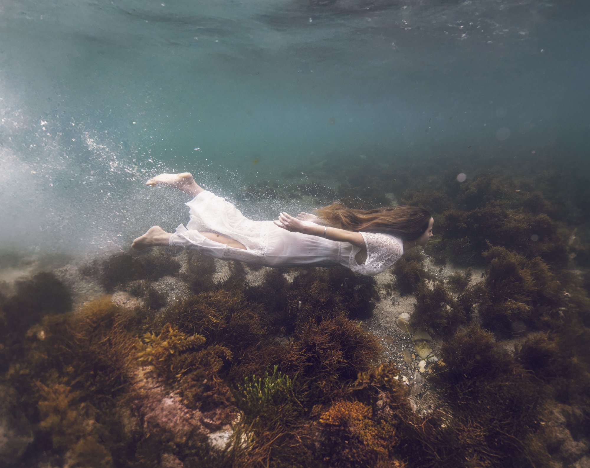 Woman swimming underwater near a coral reef, wearing a white dress with long brown hair flowing behind her.