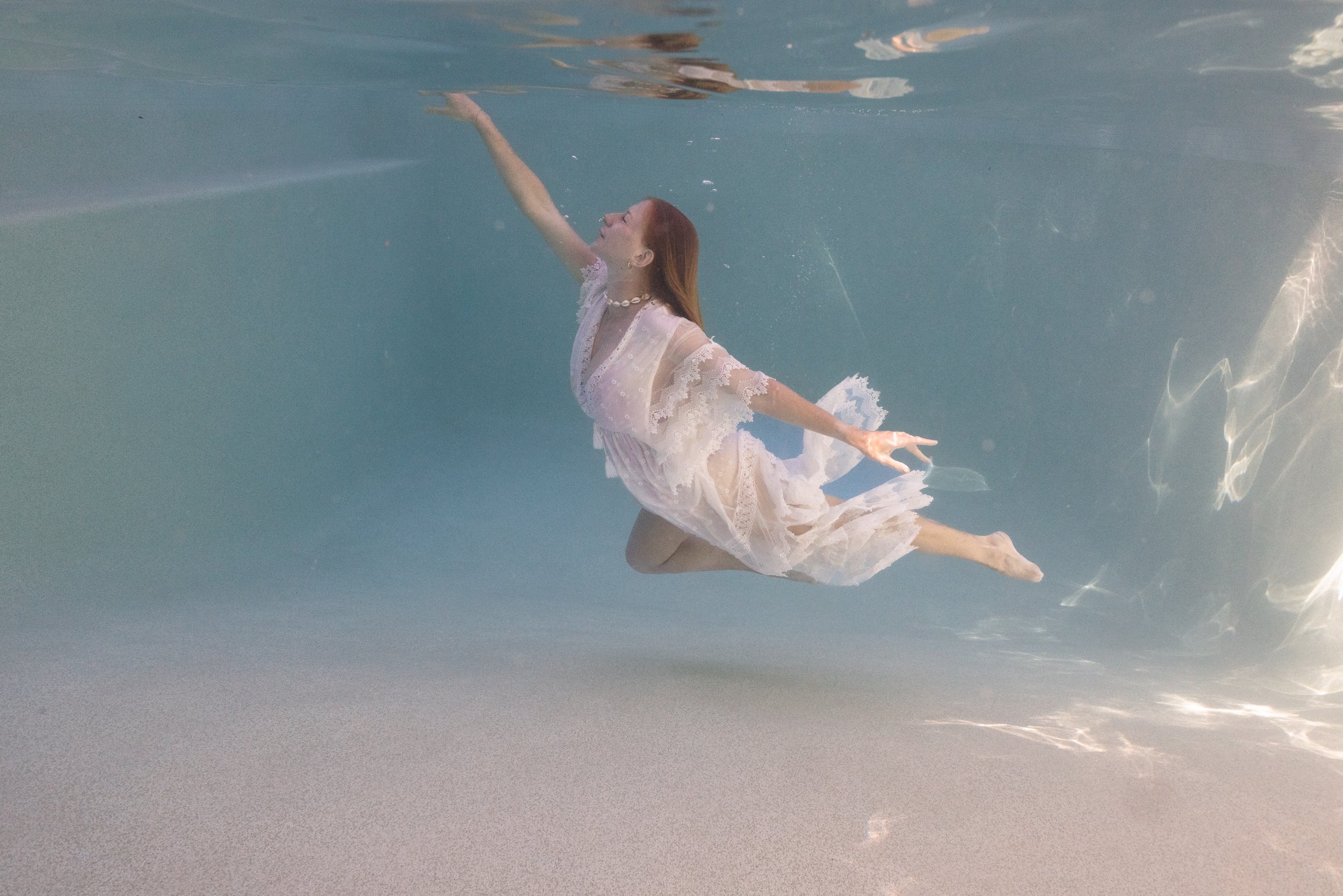 A woman with red hair swimming underwater in a white lace dress with shell necklace.
