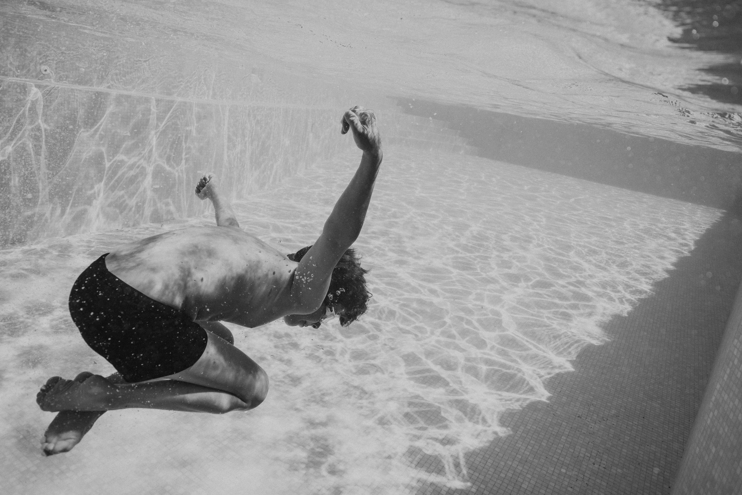 A person with long hair and sunglasses falling backwards into a swimming pool, water splashing around, wearing black swim shorts.