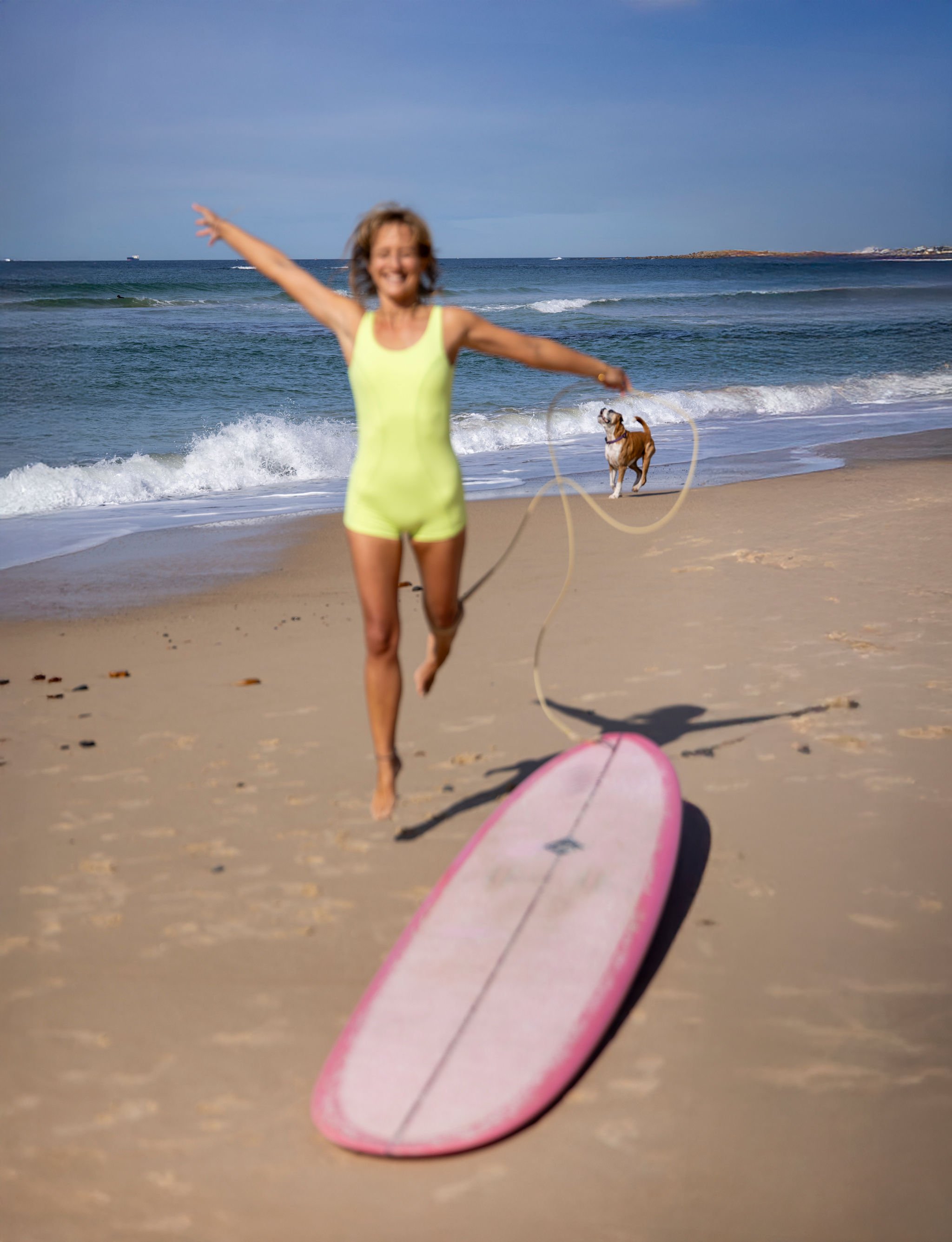 A woman in a yellow swimsuit running on the beach with a pink surfboard in the foreground, and a dog playing with a ball in the background near the shoreline.