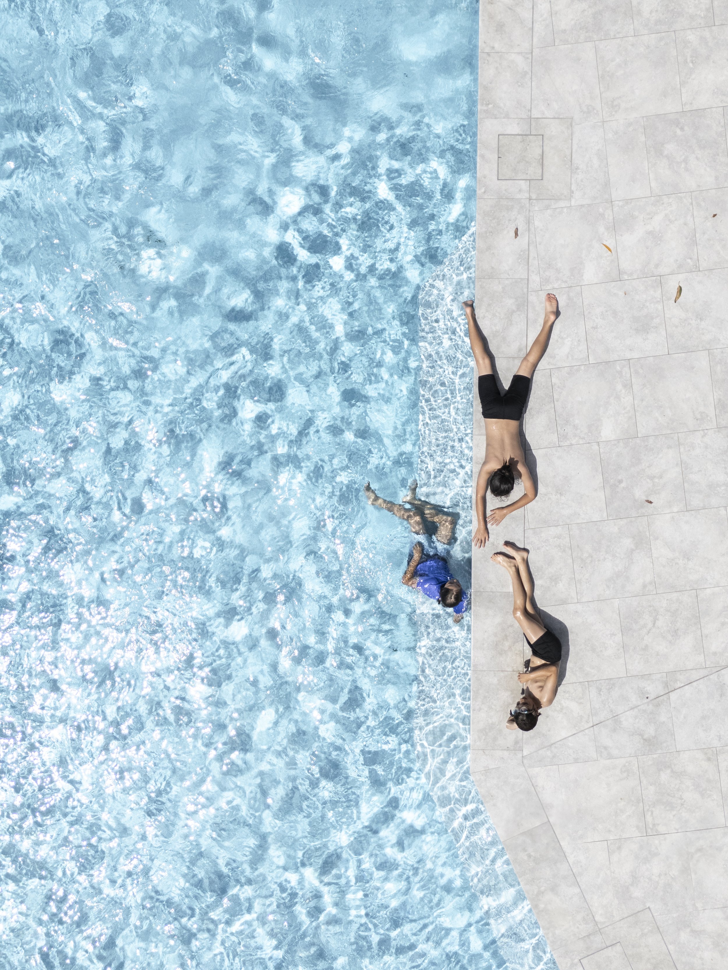 Two women and a child relaxing by the edge of a swimming pool, one woman lying on the pool deck, the child swimming in the water, and the other woman sitting on the edge.