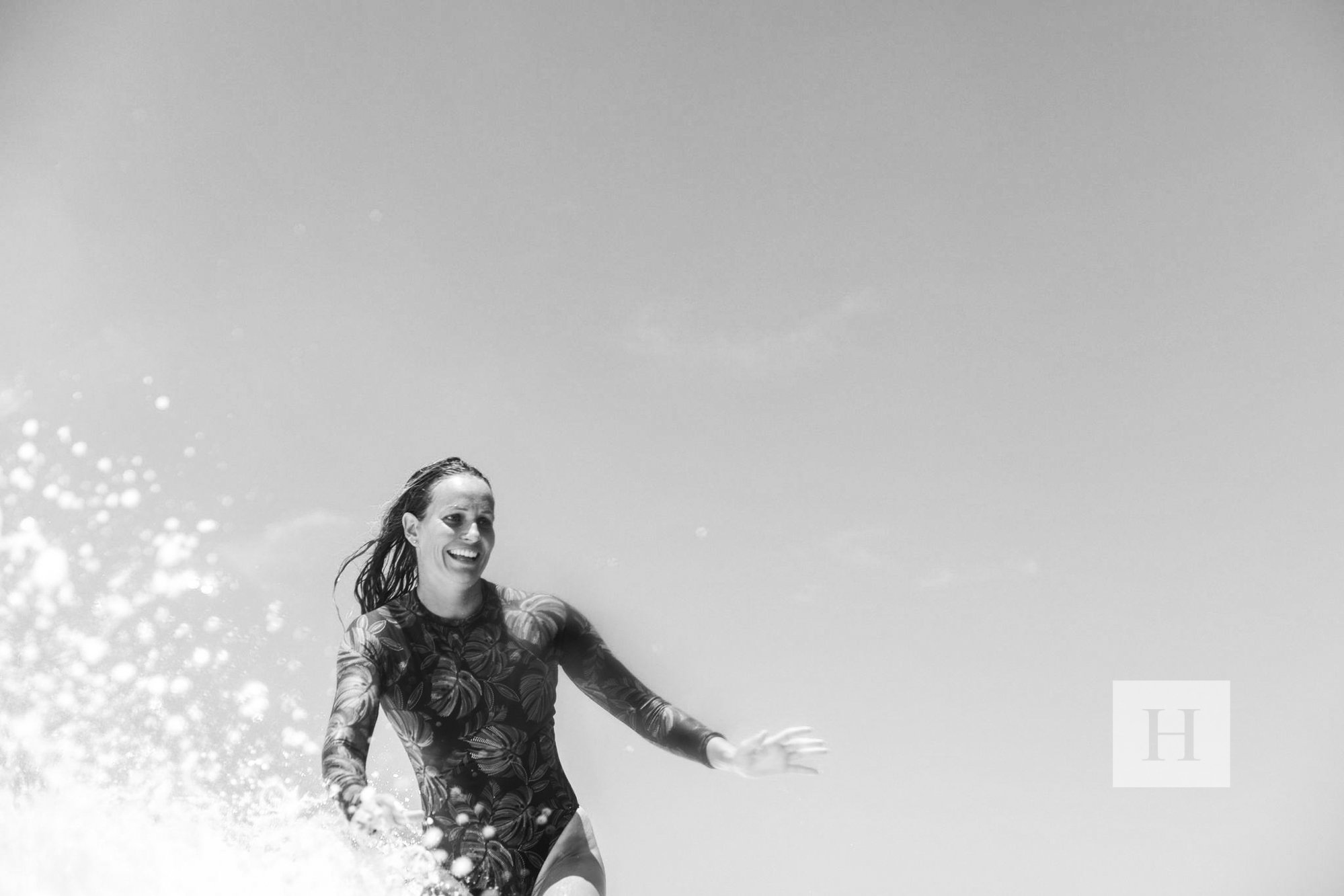 A woman in a swimsuit running in the water, smiling with waves splashing around her, under a clear sky.