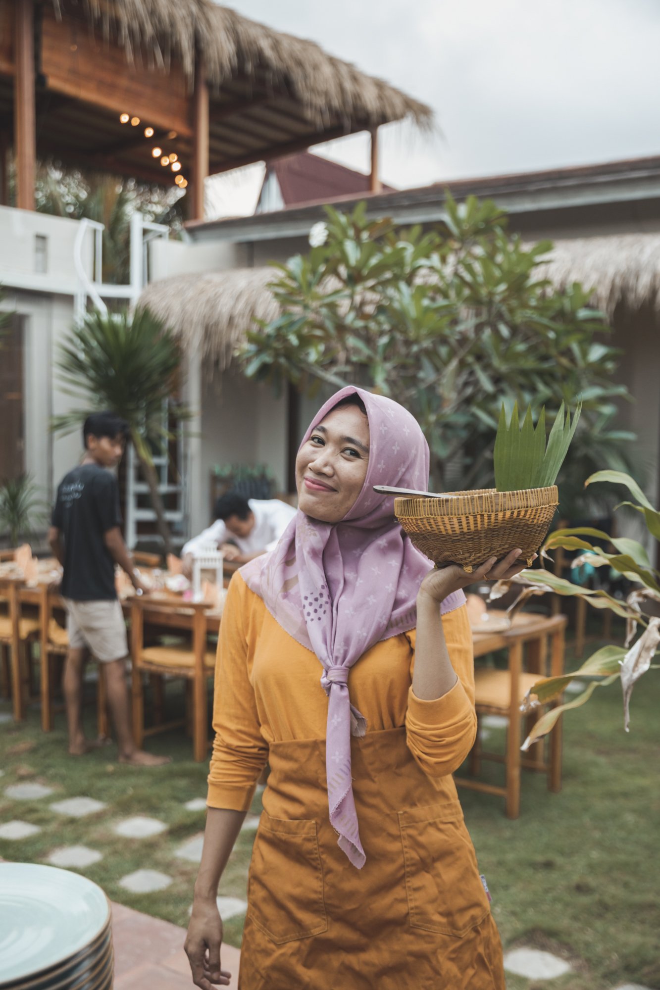 A woman wearing an orange dress and a pink headscarf holding a basket with a large green leaf, standing outdoors in a garden or patio setting with other people and tables in the background.