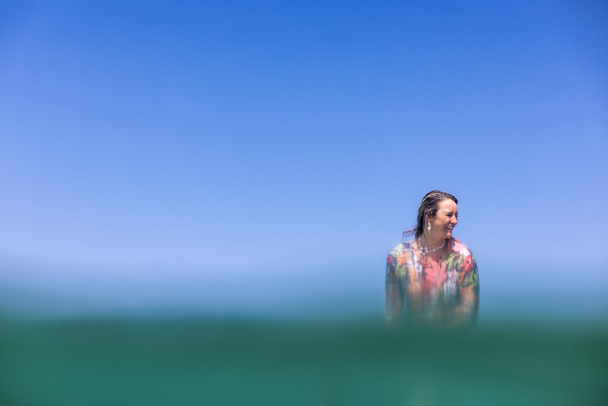 A woman with wet hair wearing a colorful swimsuit and jewelry, smiling while standing in the ocean with the clear blue sky above.