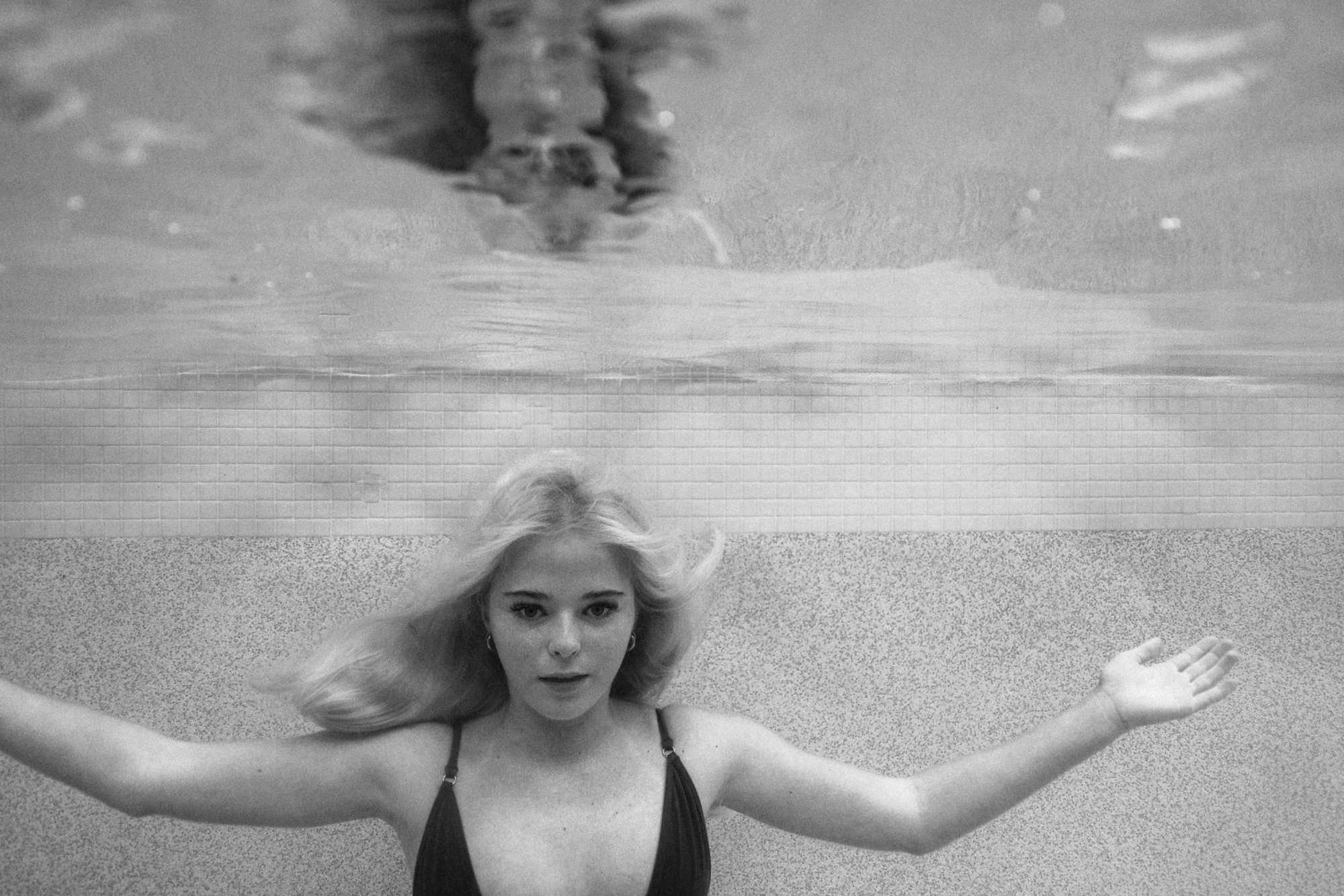 Black and white photograph of a person submerged underwater, with arms extended to the sides, and hair floating around them in a swimming pool.