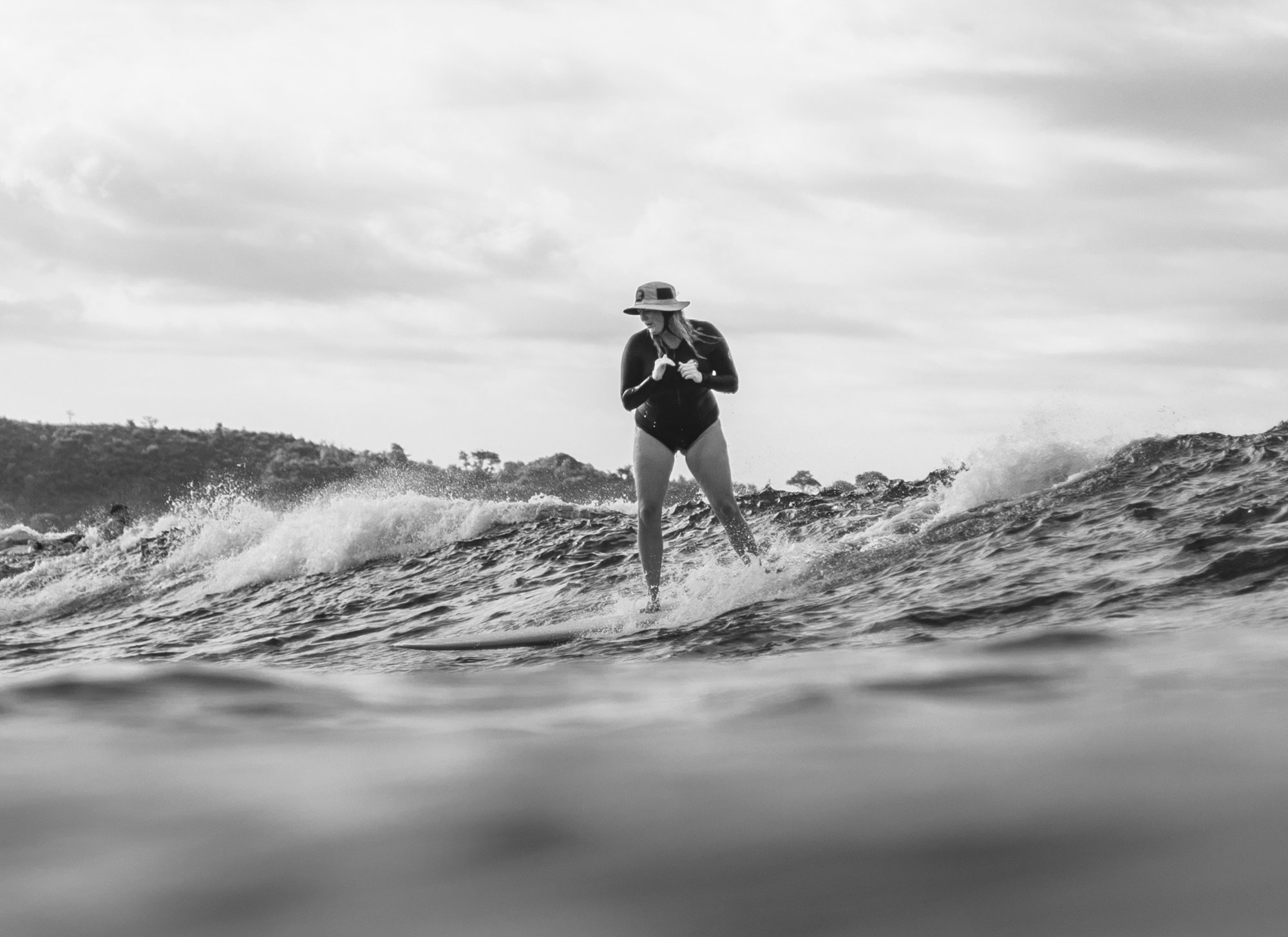A woman wearing a hat and wetsuit surfing on a wave in black and white.