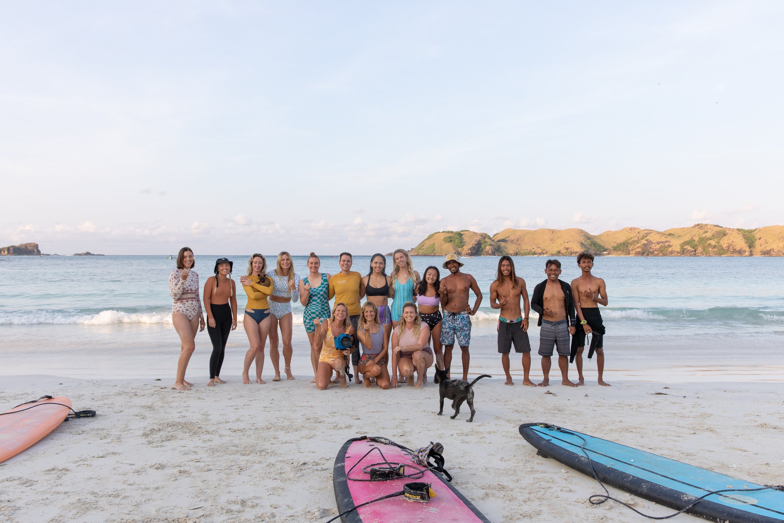 A group of people, including men and women, standing on a beach with surfboards, a dog, and ocean in the background.
