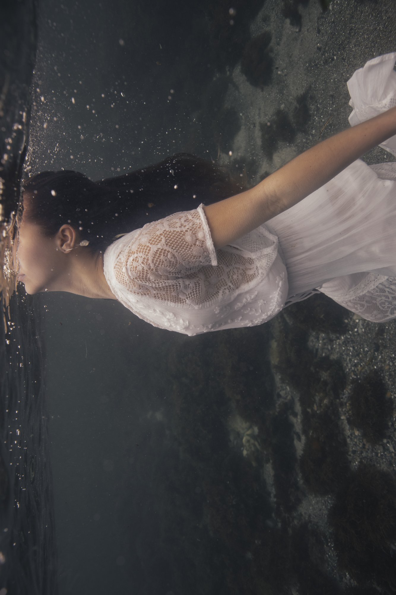 A woman in a white lace dress is underwater, facing sideways with her eyes closed, with her long black hair floating and water droplets surrounding her.