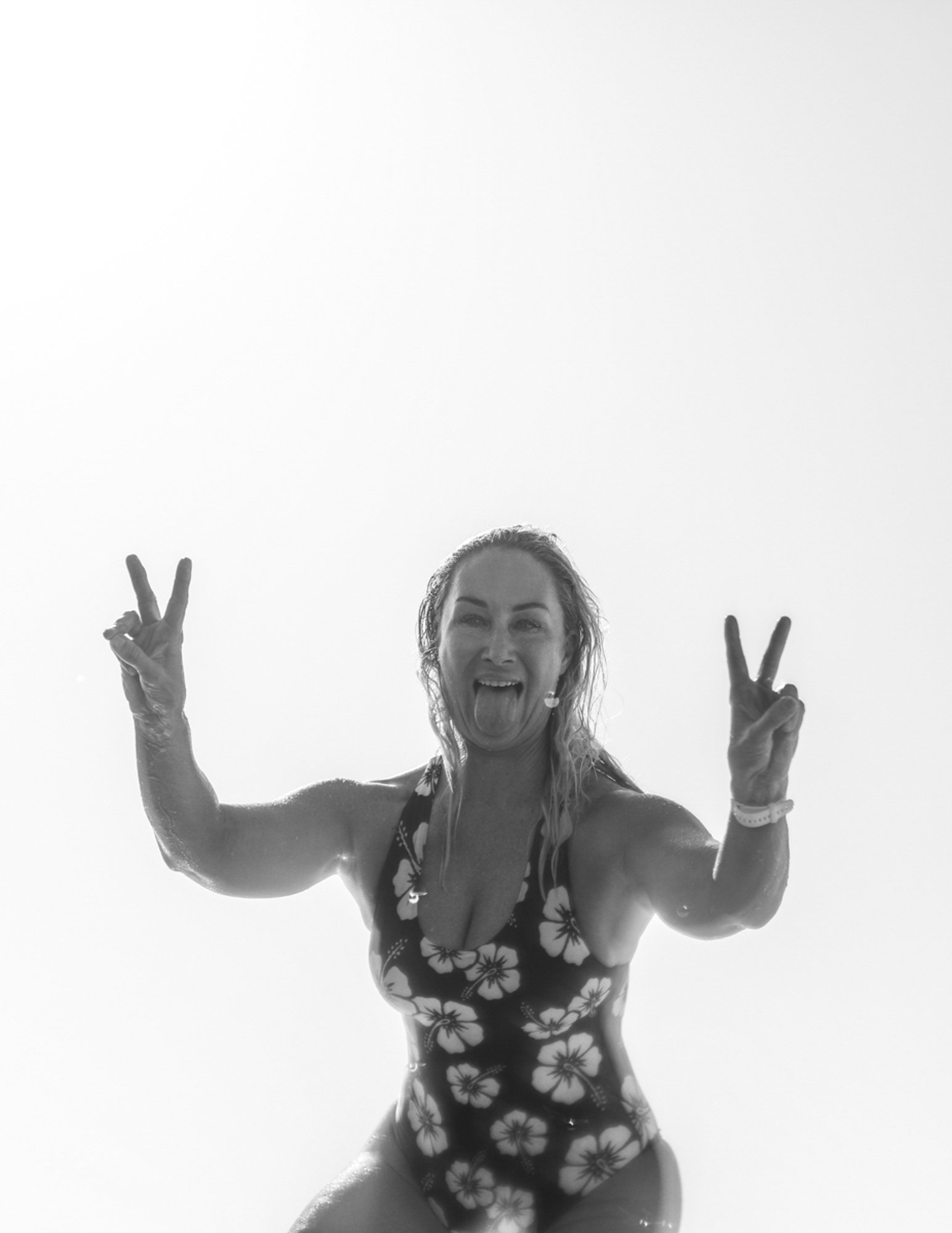 Woman in a floral swimsuit making peace signs with both hands, sticking out her tongue, smiling, and wet, against a bright background.