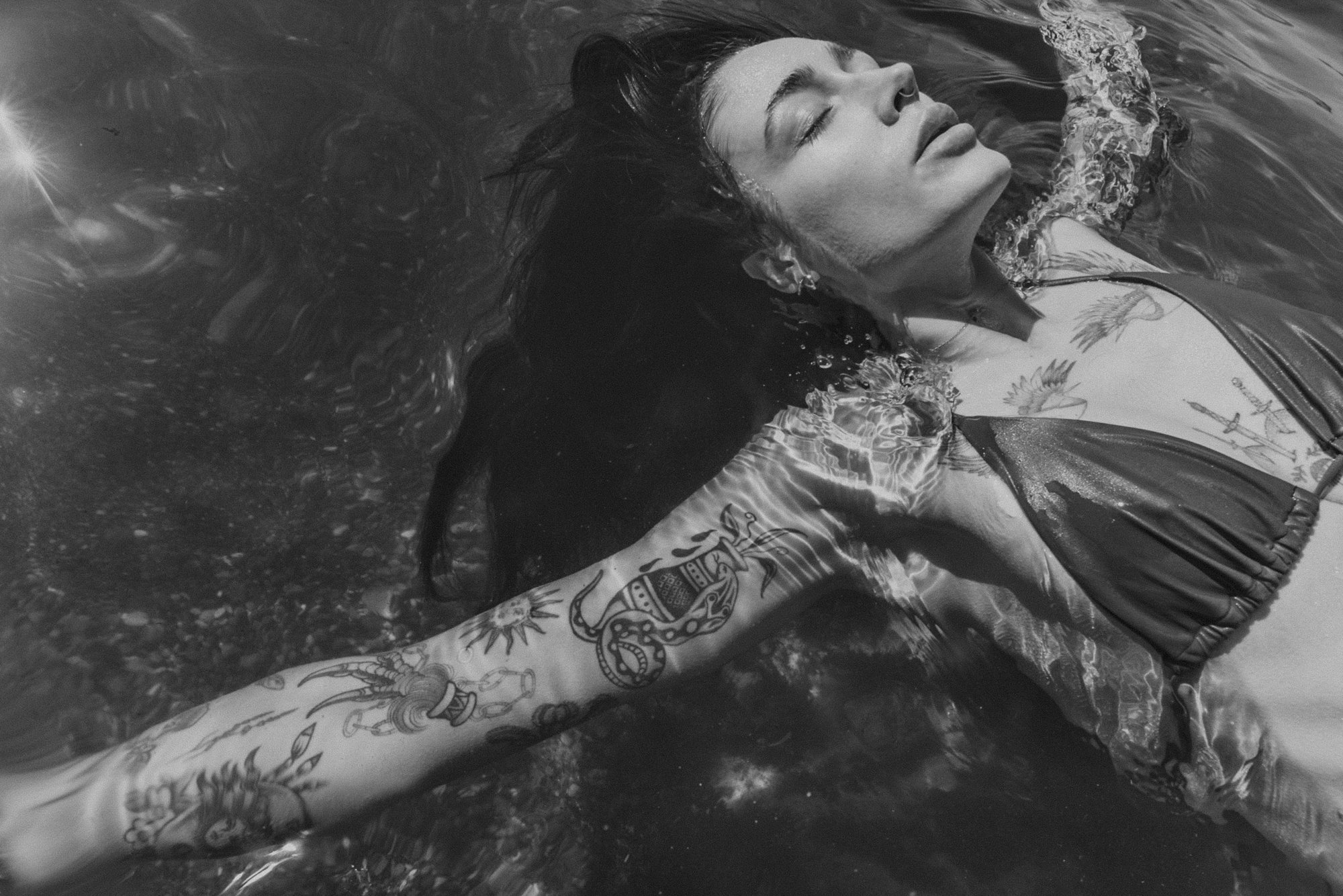Black and white photo of a person floating in water, wearing a bikini, with tattoos on the arm and chest. The person's eyes are closed, and their hair is spread out in the water.