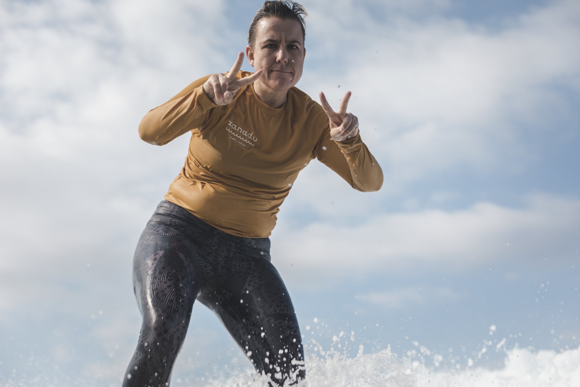 Woman wearing a tan long-sleeve shirt and black patterned leggings making peace signs with both hands while splashing in water against a cloudy sky background.
