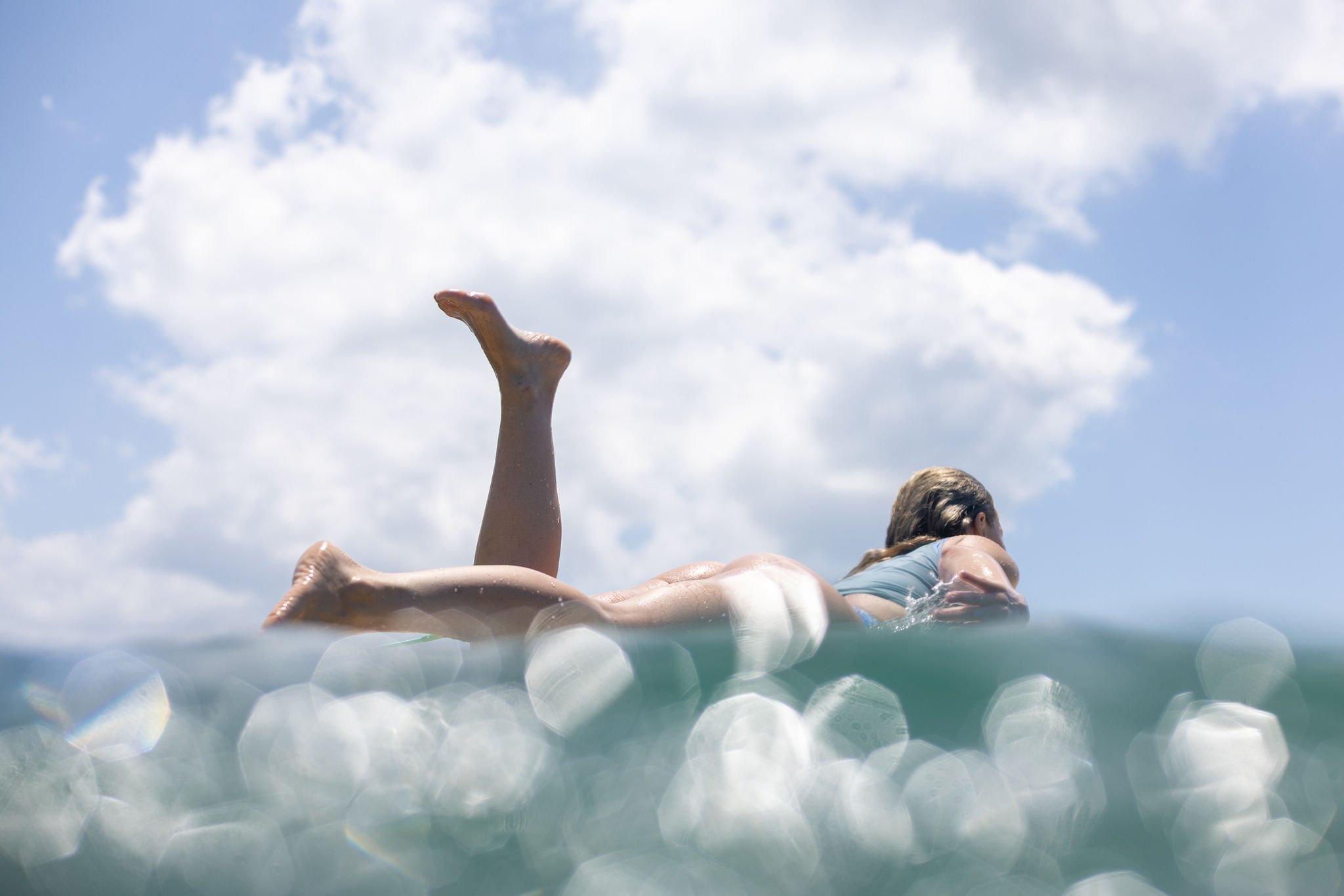 A woman swimming or floating in the ocean during the daytime, with her body partially submerged and her face turned away, under a partly cloudy sky.