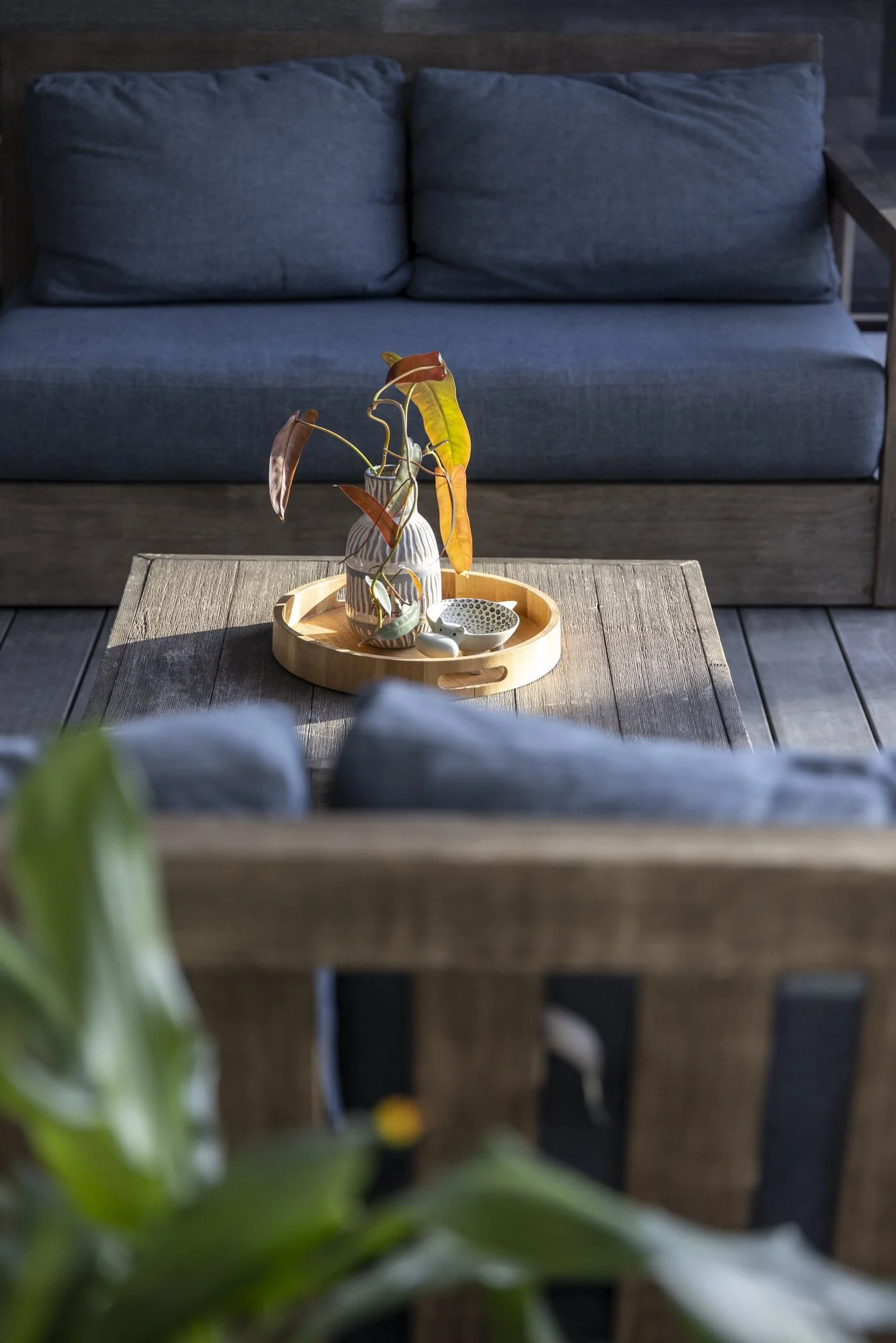 View of a cozy indoor space with a dark blue fabric sofa in the background, a wooden coffee table in the foreground with a round wooden tray holding a decorative vase with brown and yellow leaves, and two small decorative bowls, one patterned, on the