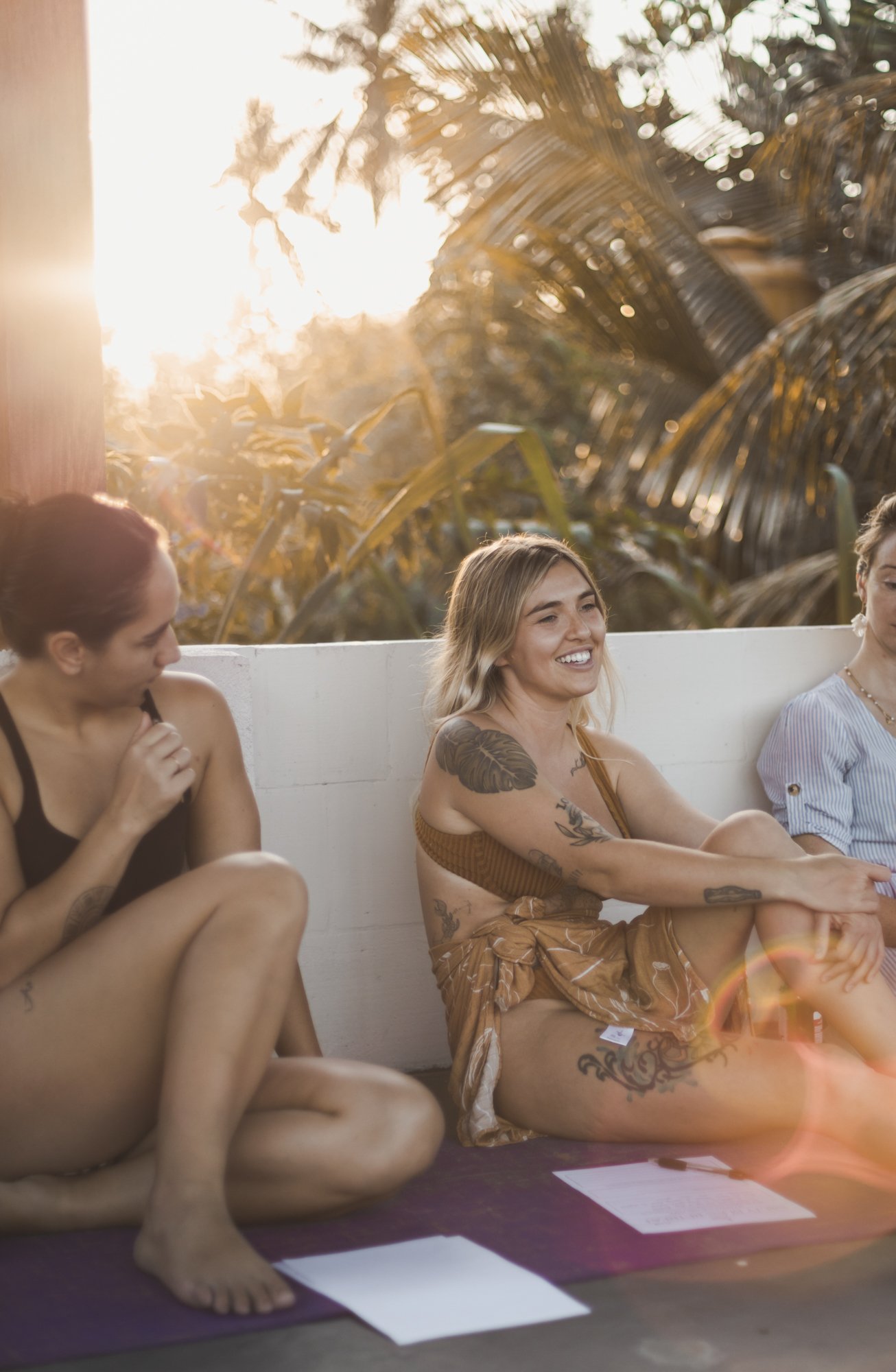 Three women sitting on mats, smiling and talking outdoors during sunset, with tropical plants in the background.