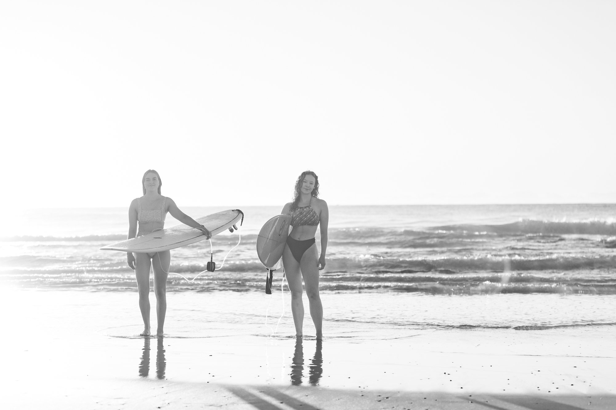 Two women in swimsuits holding surfboards and standing in shallow ocean water at the beach during sunset.