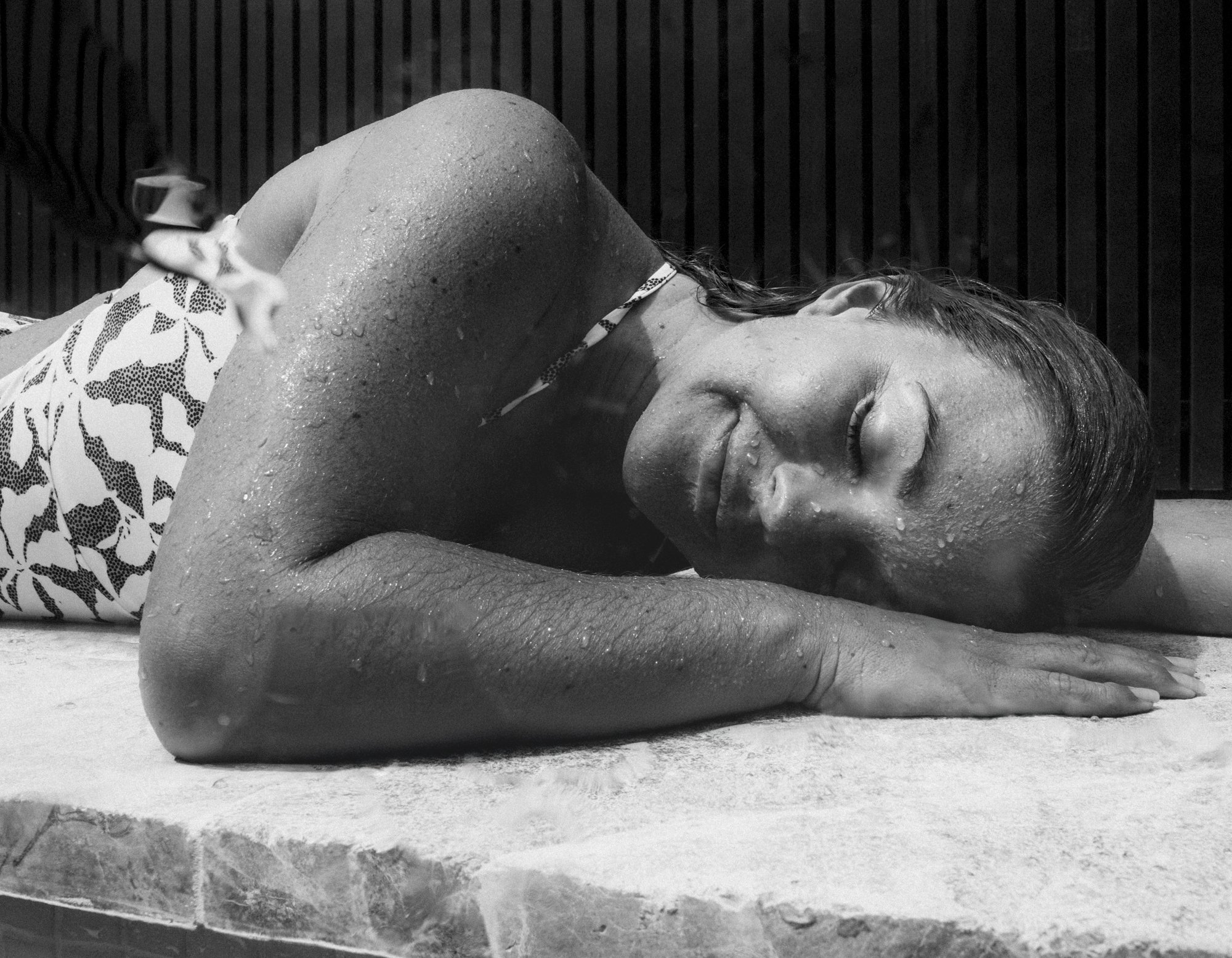 A woman with wet hair and water droplets on her face and arm, lying on her side with her eyes closed, on a surface near a wooden wall.