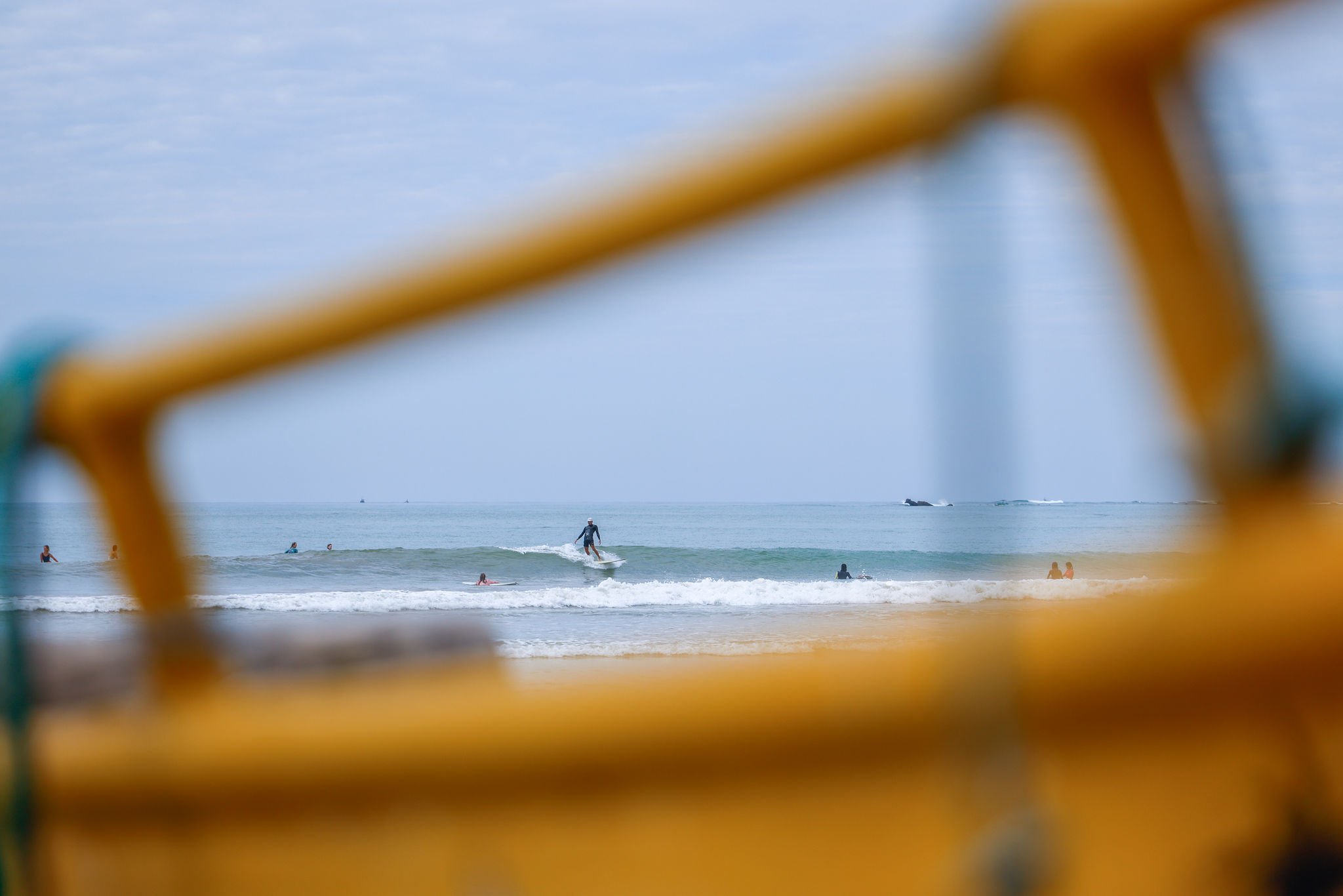 View of a beach scene with people swimming and surfing, seen through a blurry yellow safety fence.