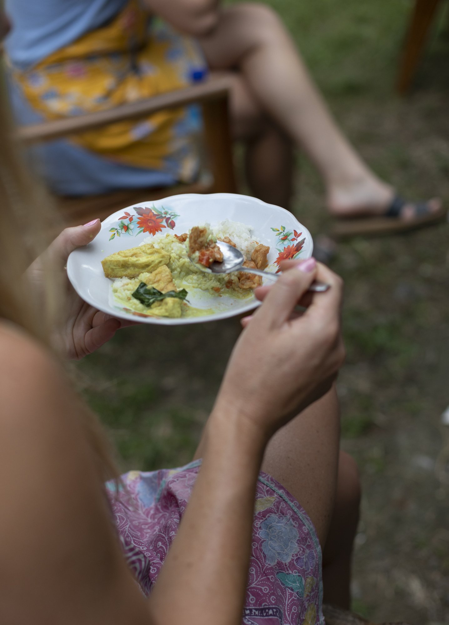 A woman holding a floral ceramic plate with rice and curry, sitting outdoors on a wooden chair, with someone in the background also seated outdoors.