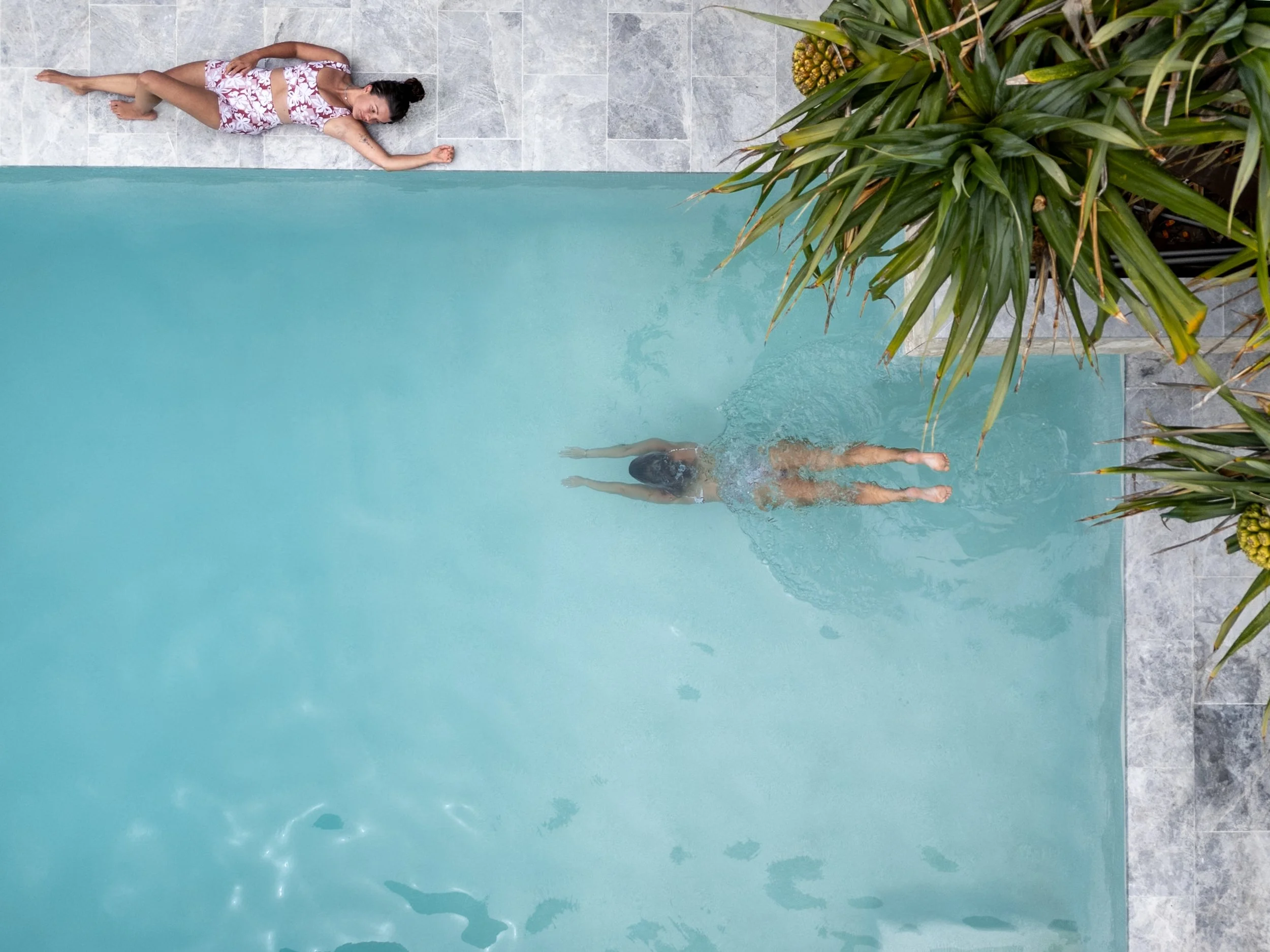A woman in a swimsuit is diving into a swimming pool, with tropical plants and a woman lying on the poolside.