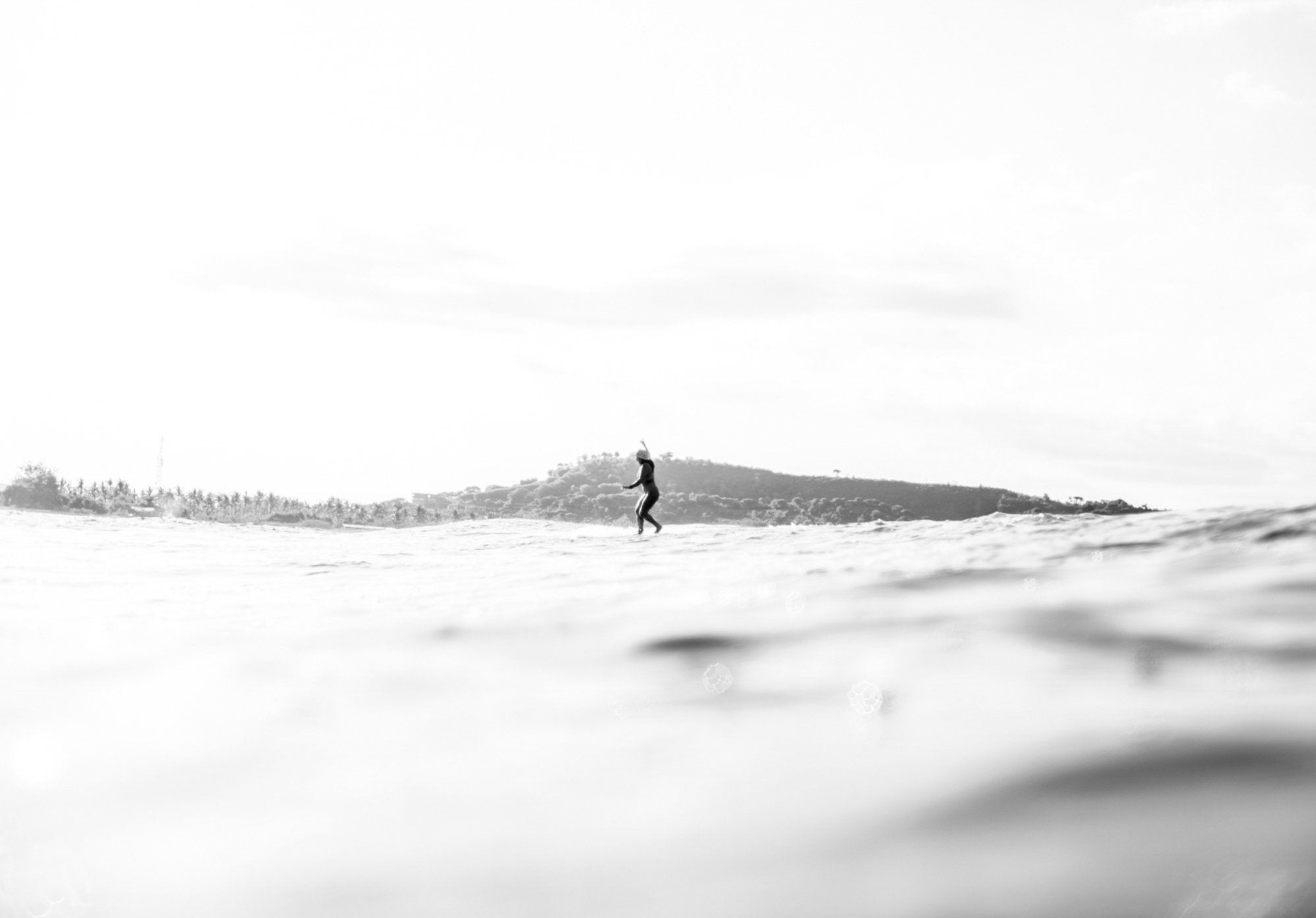 A person surfing on a wave in the ocean, with a distant shoreline and hills in the background, in black and white.