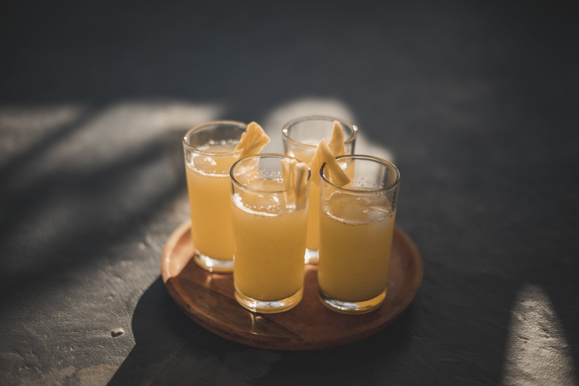 Four glasses of yellow beverage with lemon slices on a small wooden tray, placed on a dark surface with soft lighting.