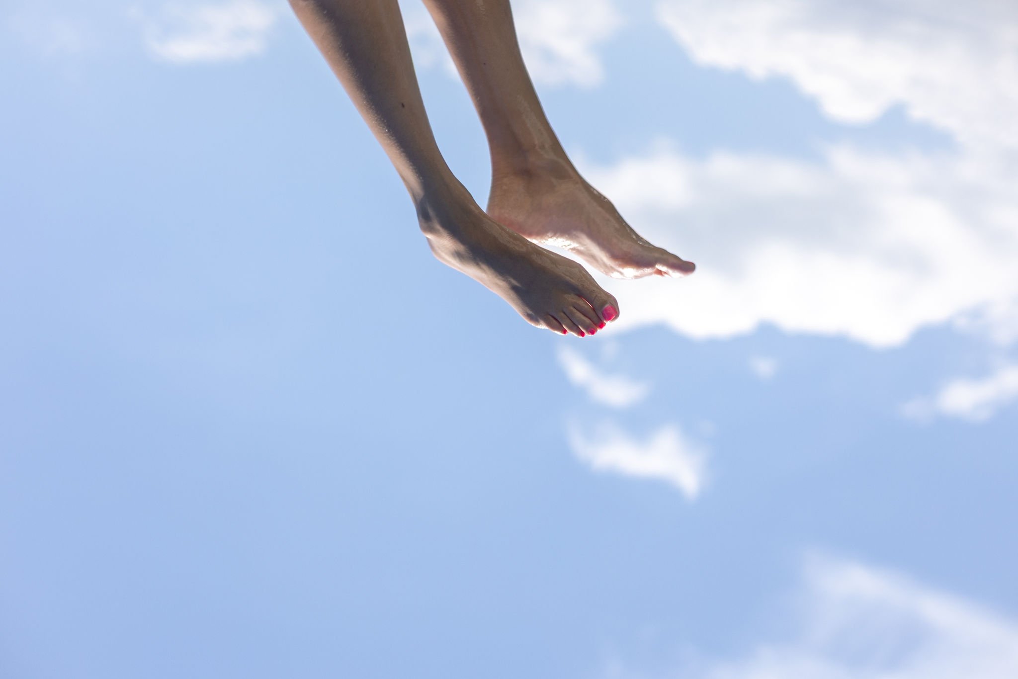 Close-up of a person's legs and feet with pink nail polish, hanging upside down against a bright blue sky with scattered white clouds.