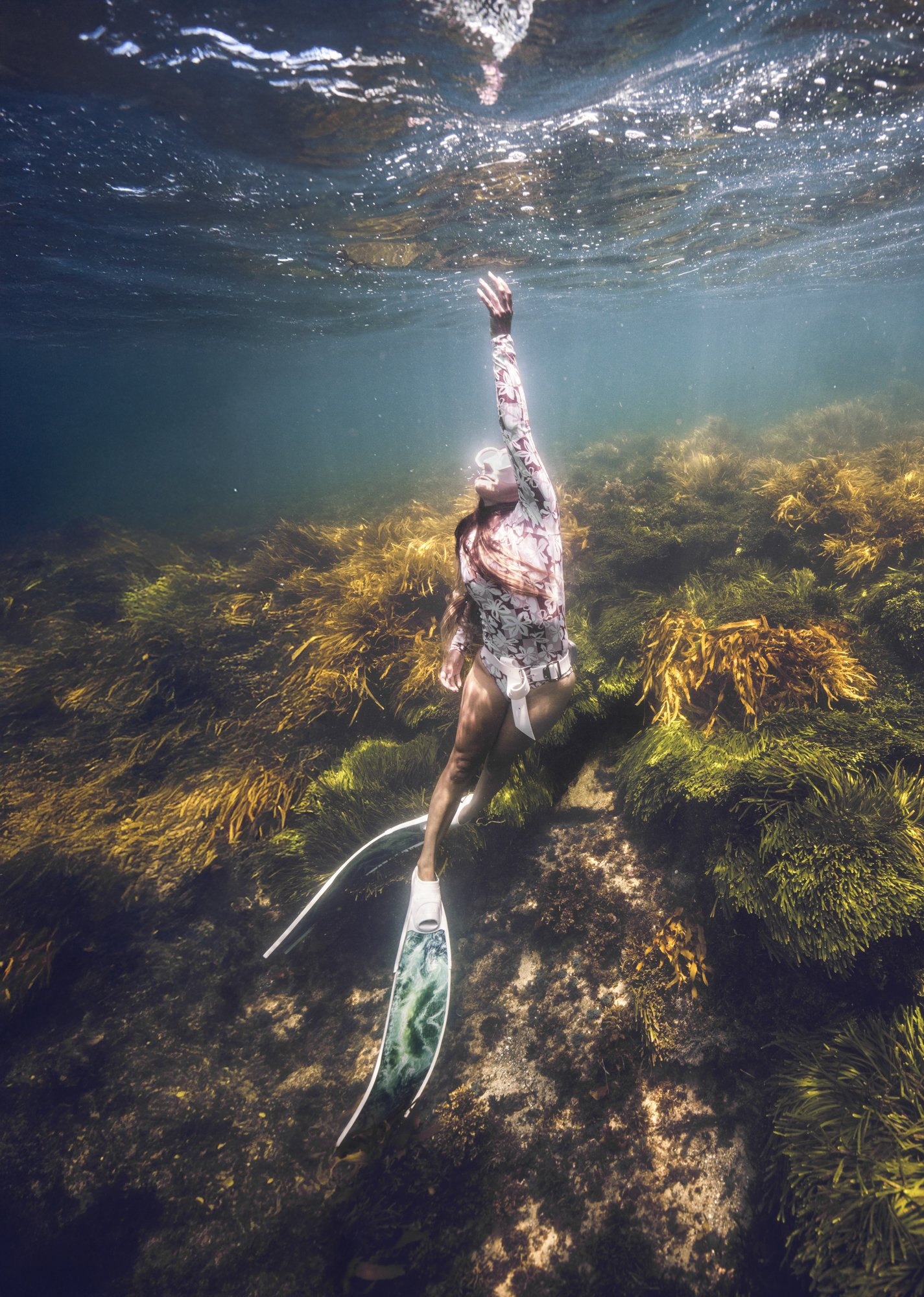 Woman in floral shirt and shorts swimming underwater near coral reef with snorkeling fins.