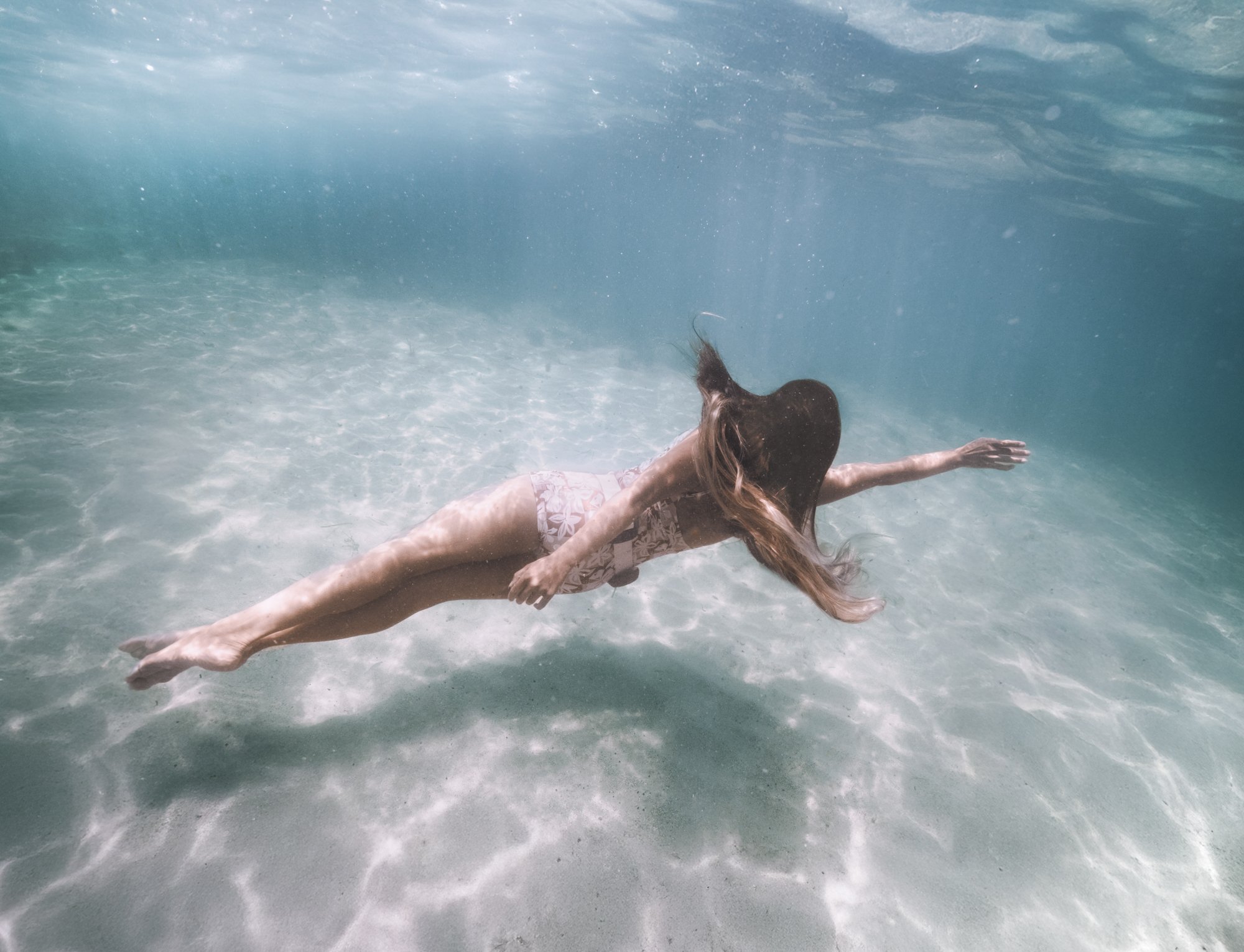 A woman swimming underwater in clear ocean water with sunlight streaming down.