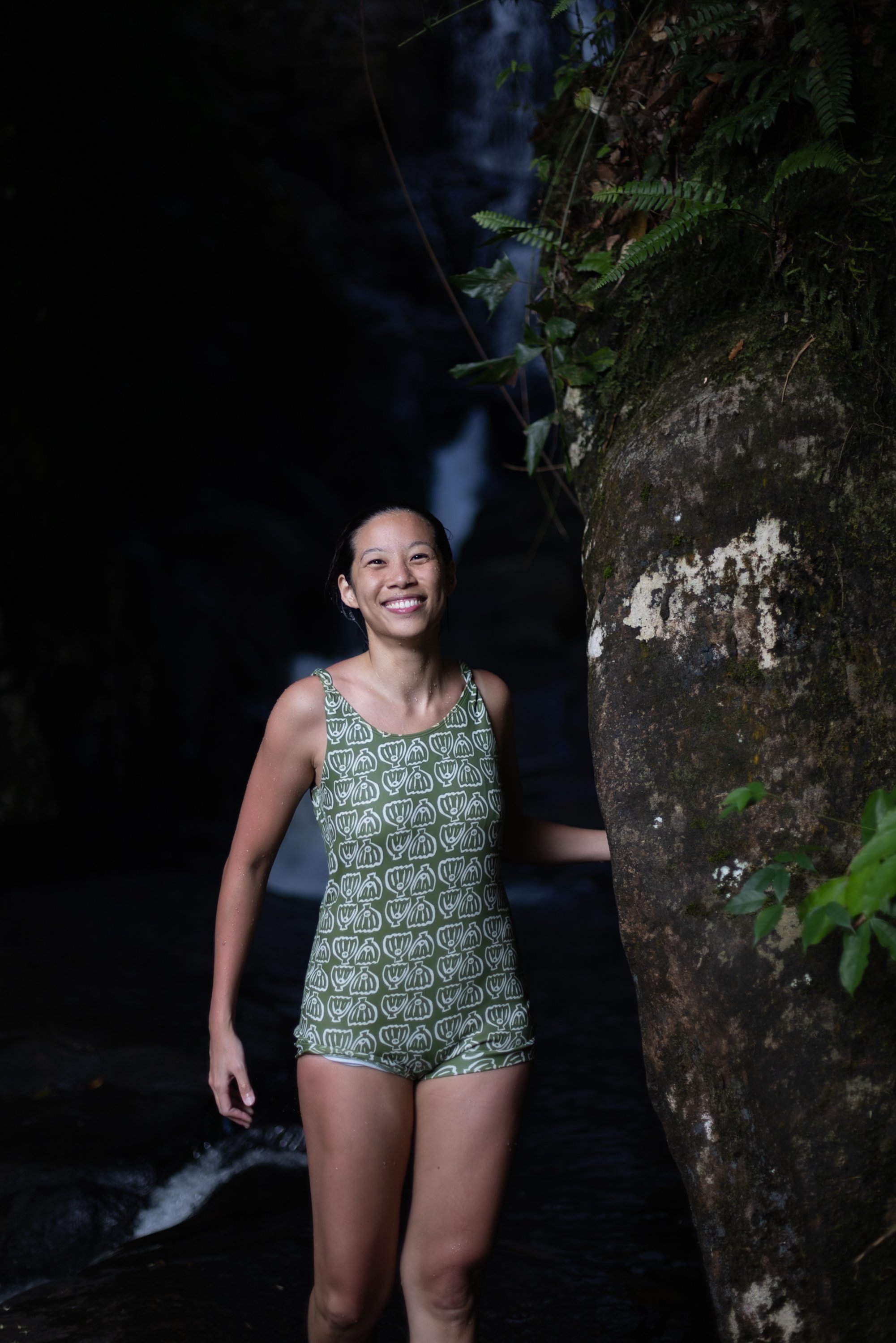 A woman smiling and standing in a shallow stream in a lush, green forest with a waterfall in the background.