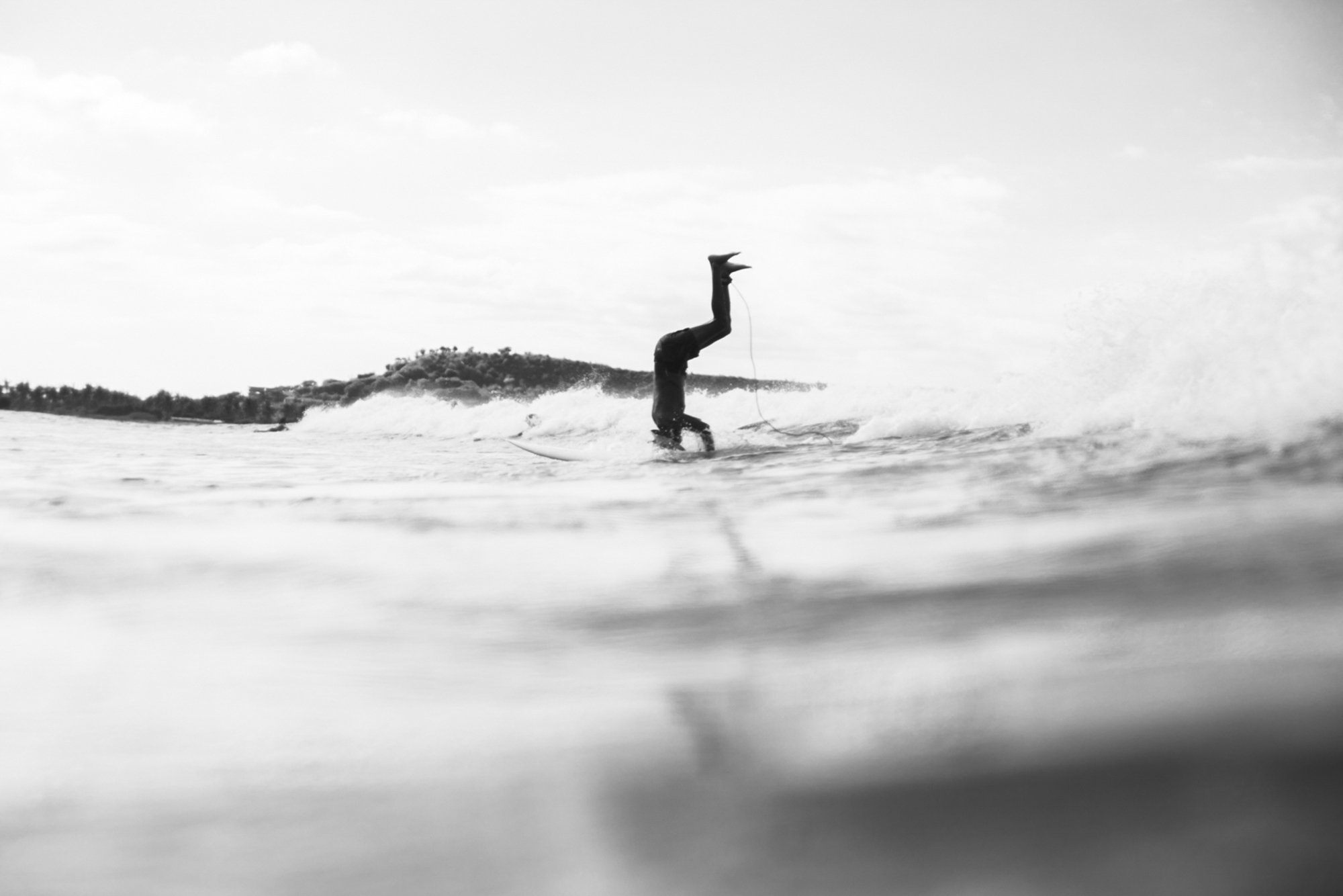 A person in a wetsuit doing a handstand on a surfboard in the ocean, black and white photo with a distant shoreline and cloudy sky