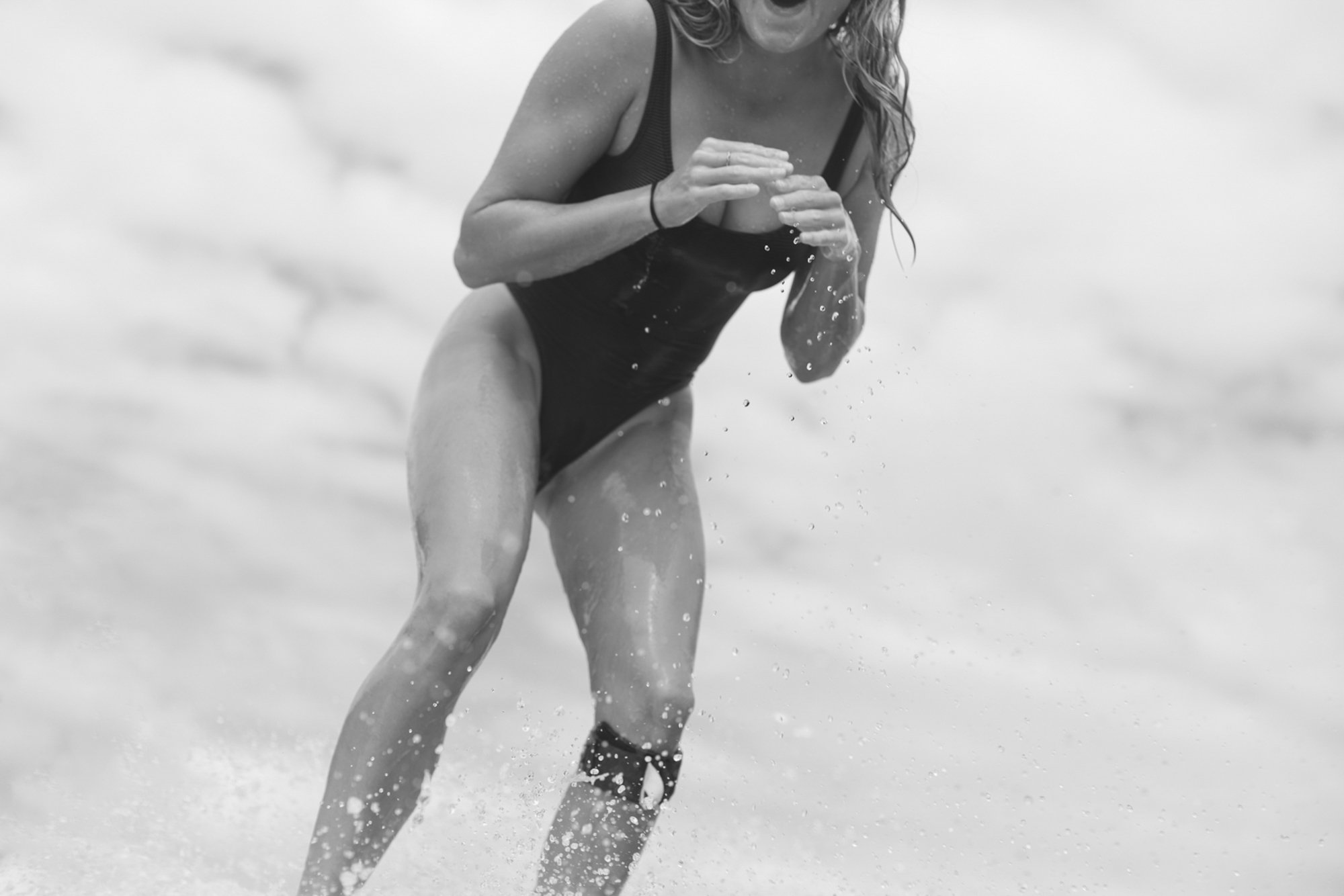 A woman in a black swimsuit running through the water at the beach, with water splashing around her.