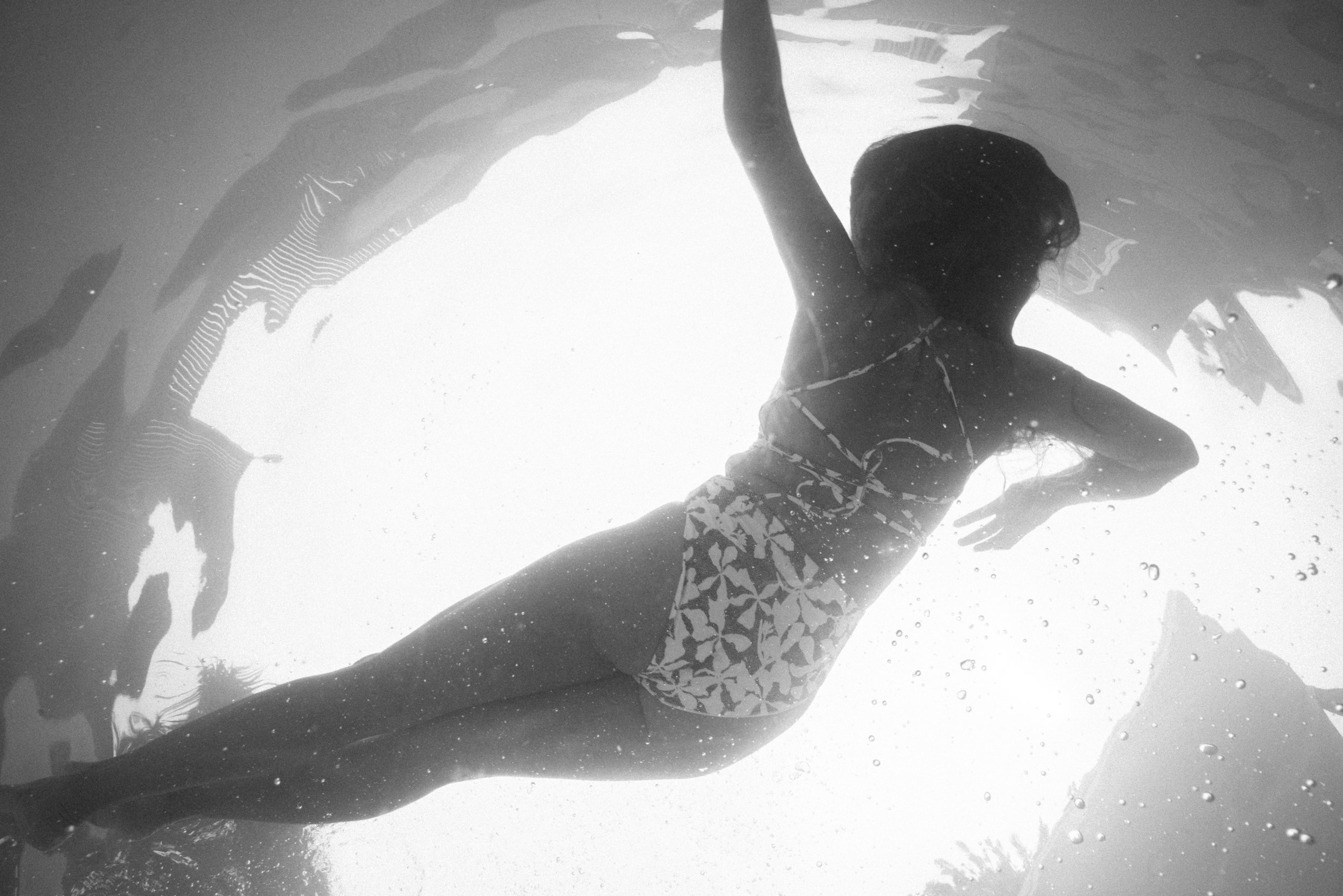 A woman swimming underwater wearing a patterned bikini with her arm raised, seen from below.