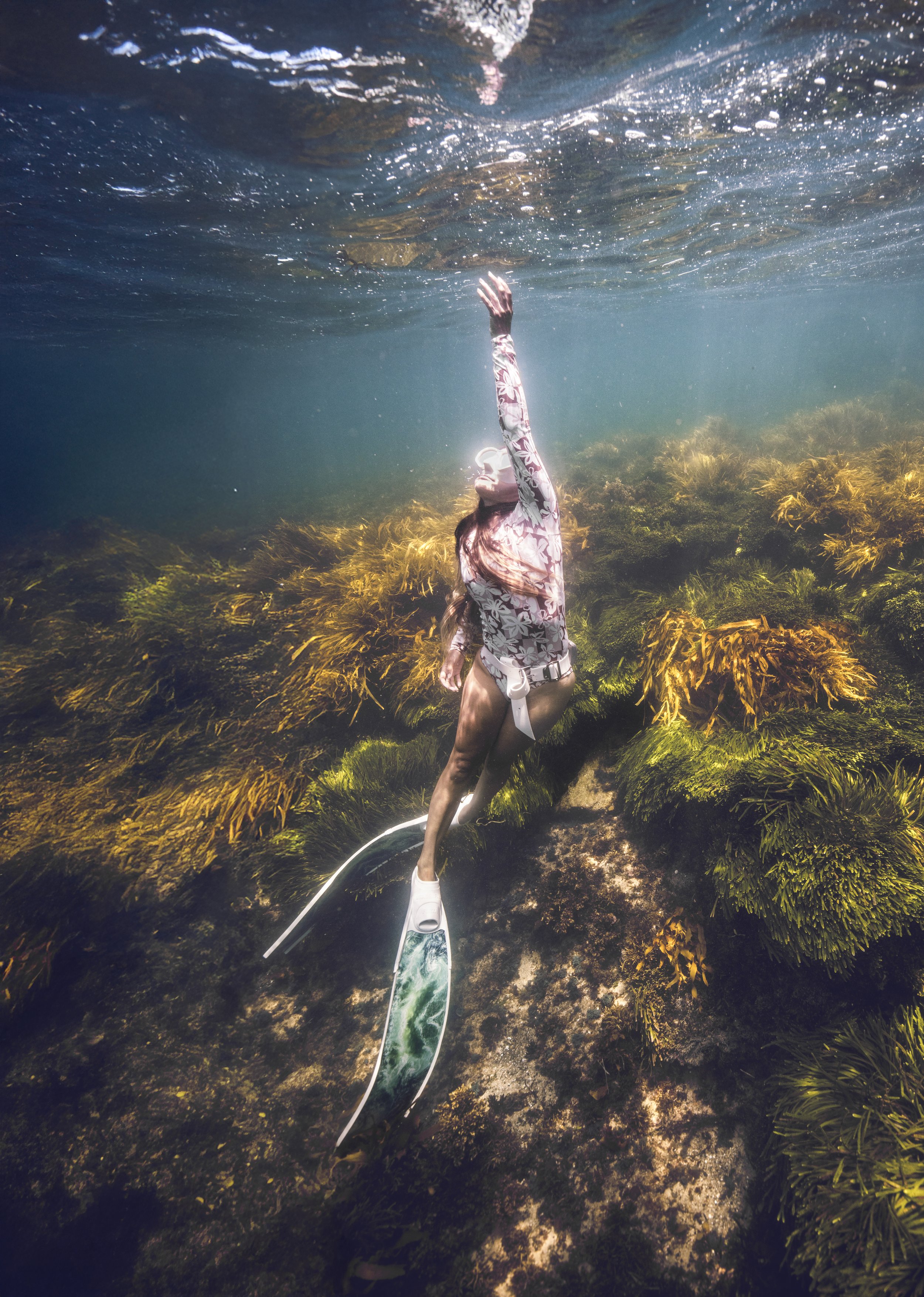 Woman snorkeling underwater among seaweed and rocks, reaching upward.