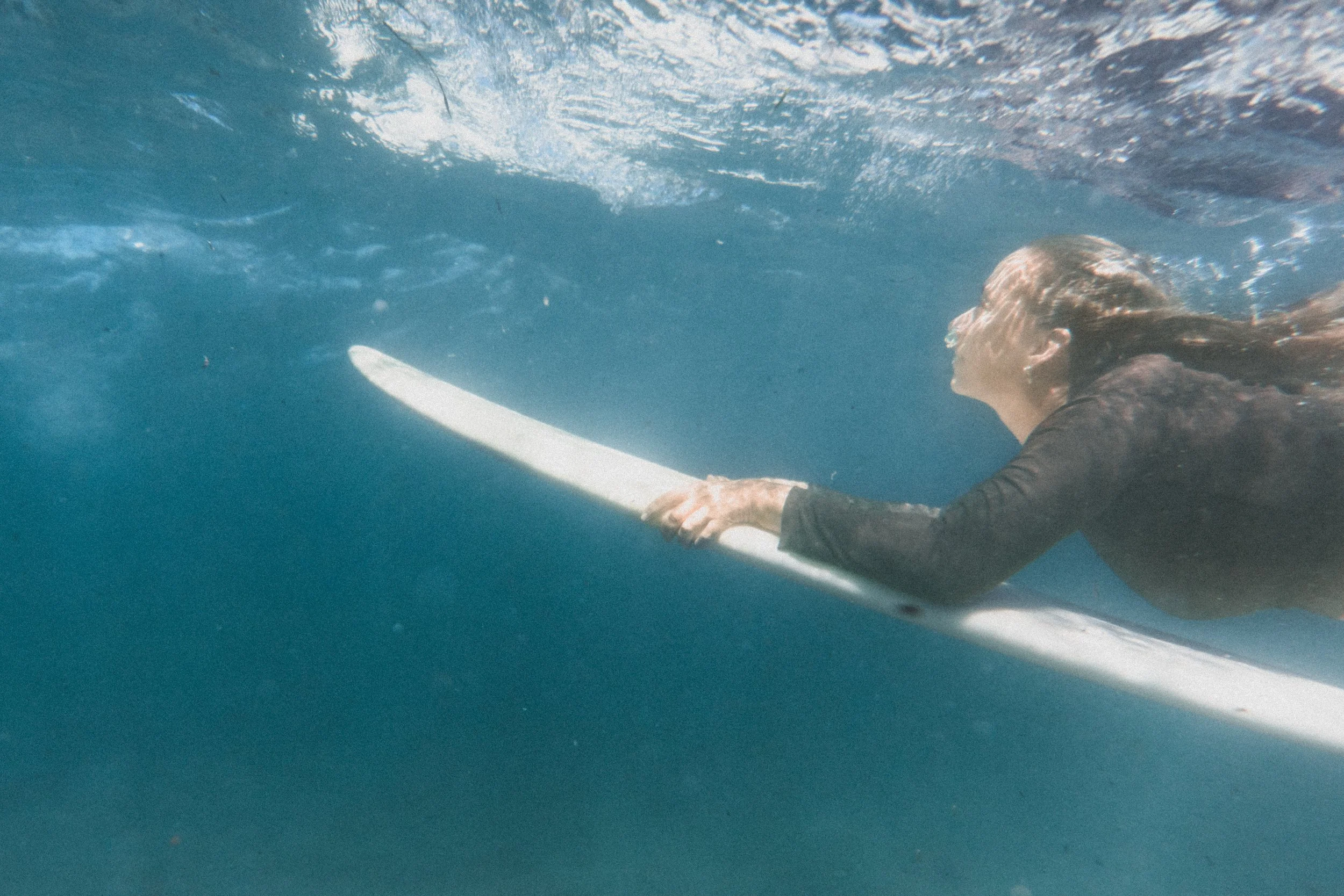 A person swimming underwater, holding a white paddle, wearing a black wetsuit, with long hair flowing behind them and the surface of the water above.