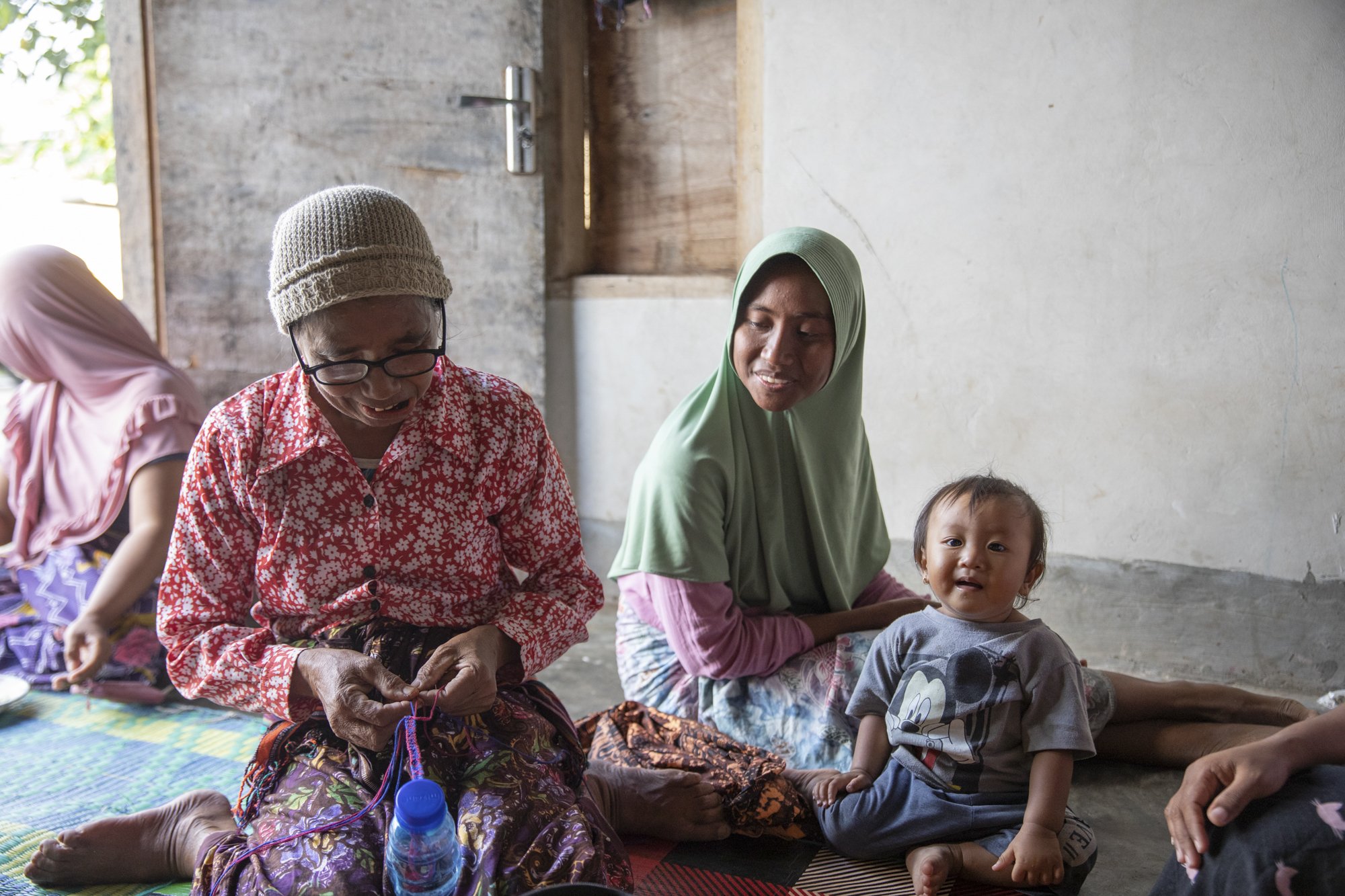 A group of women and children sitting on the floor in a simple room, interacting and smiling.