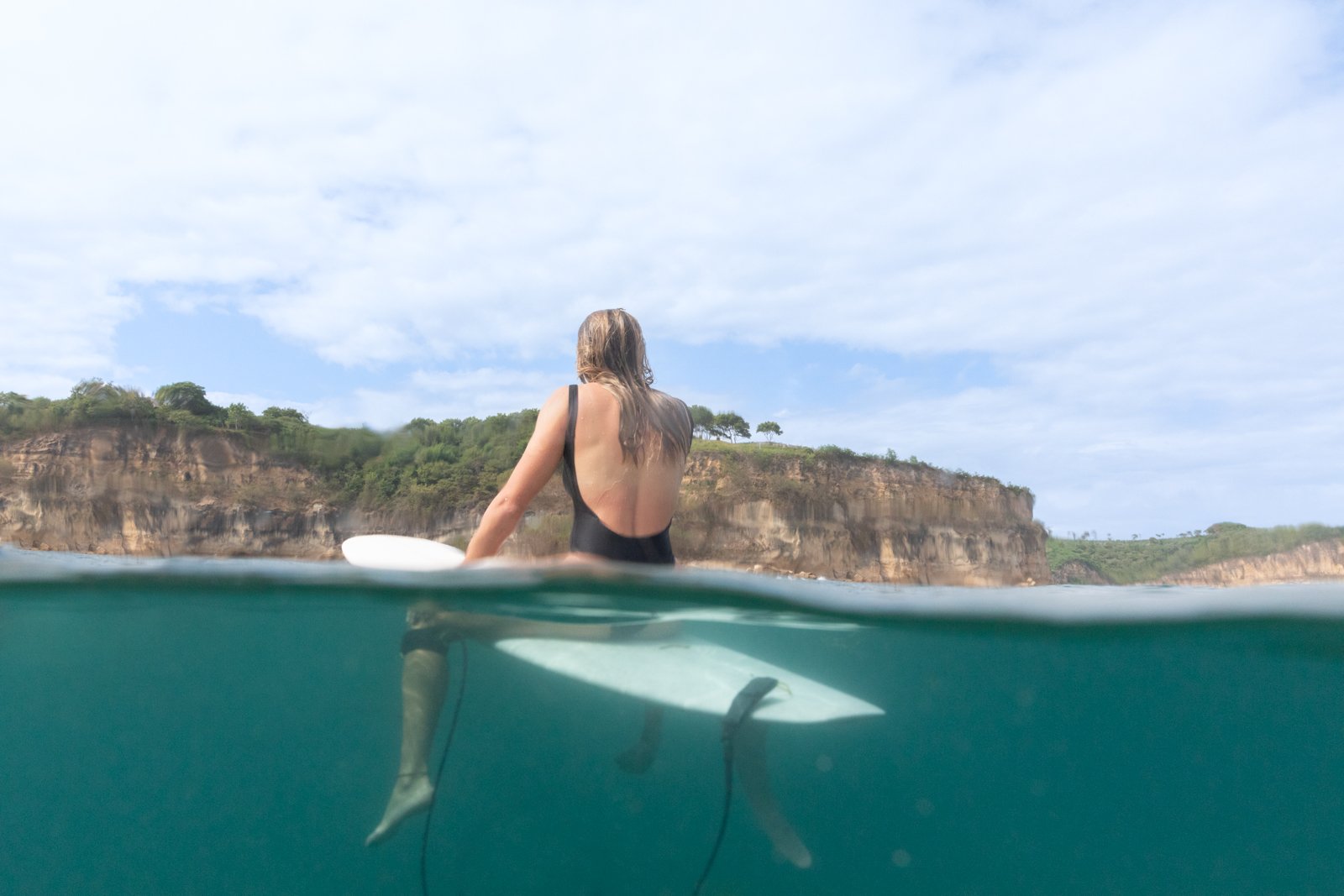 A woman in a black swimsuit sitting on a surfboard in the water, with a coastal cliff and partly cloudy sky in the background.