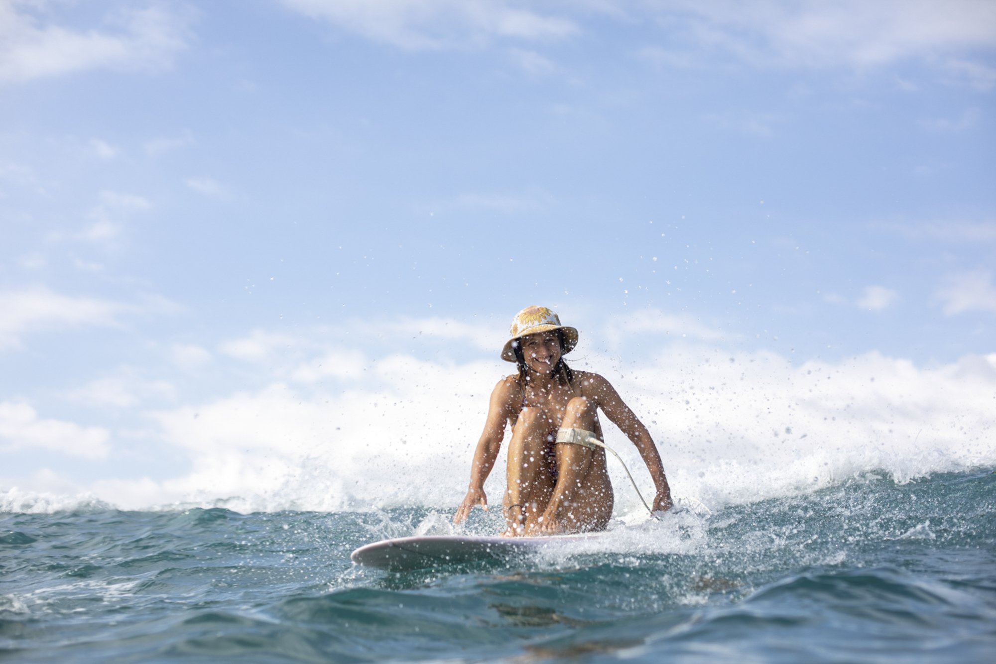 Young woman surfing on the ocean, wearing a sun hat and smiling, with blue sky above.