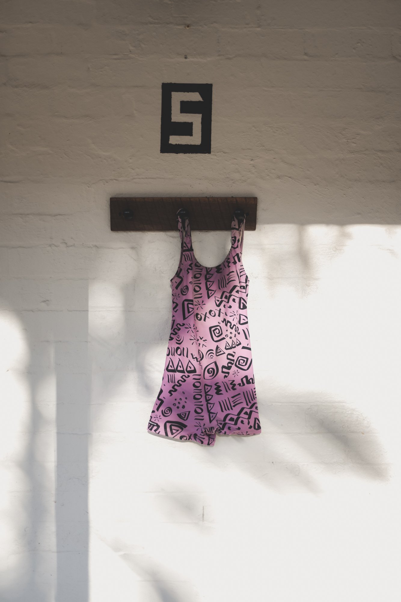 A pink dress with black abstract patterns hanging on a wooden peg rack against a white brick wall, with shadows cast on the wall and a black square with the number 5 painted above.