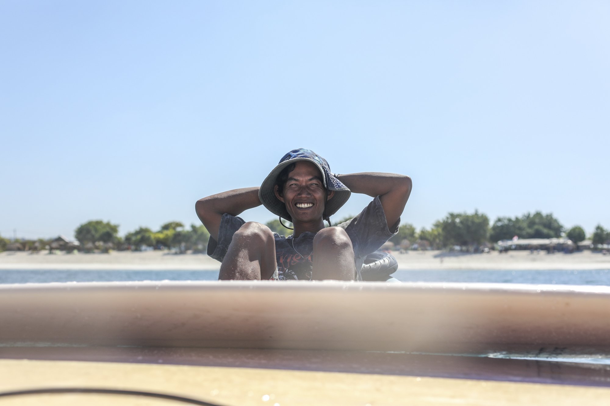 A man sitting on a surfboard at the beach, smiling and holding a wide-brimmed blue hat, with a sandy shoreline, trees, and a clear blue sky in the background.