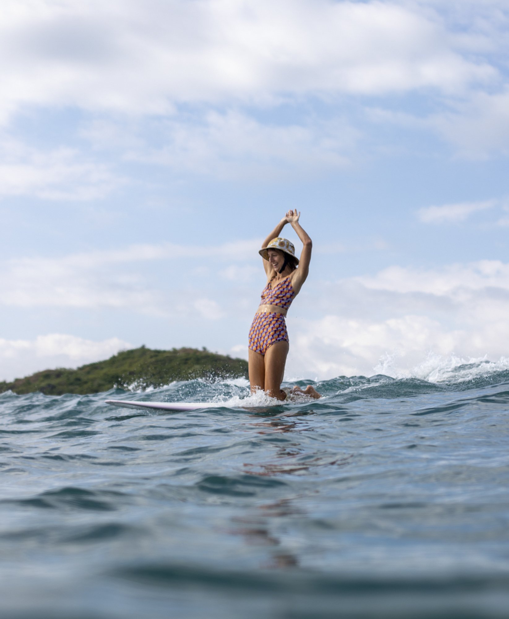 Woman in a patterned swimsuit and hat standing on a surfboard, smiling with arms raised, surfing on calm ocean waves with a small island in the background under a partly cloudy sky.