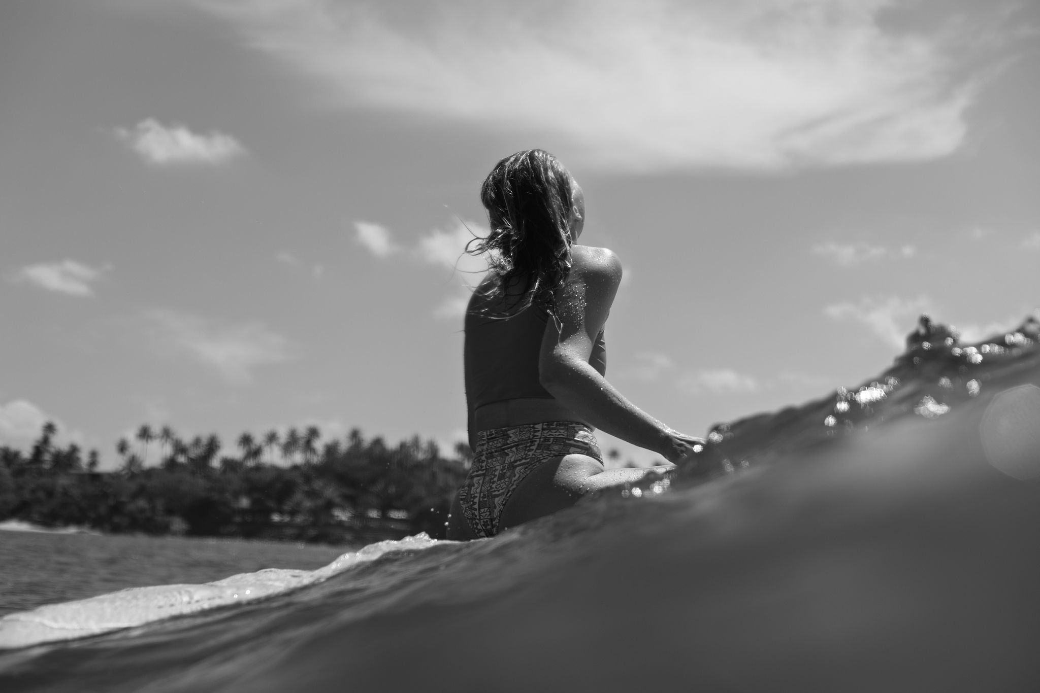 A girl sitting on a surfboard in the ocean, with palm trees in the background, under a partly cloudy sky.