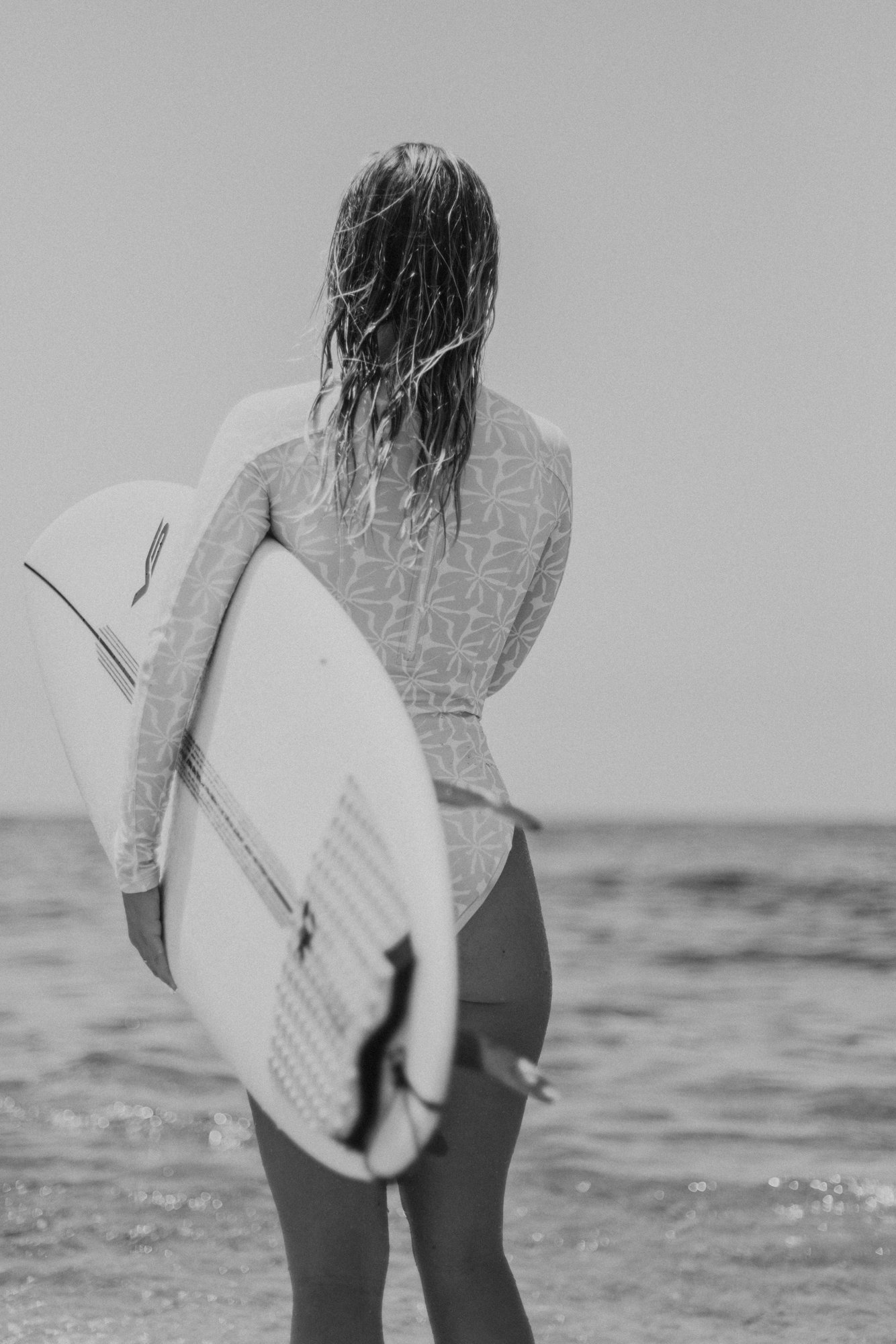 Black and white photo of a person wearing a patterned rash guard, holding a surfboard, looking at the ocean.