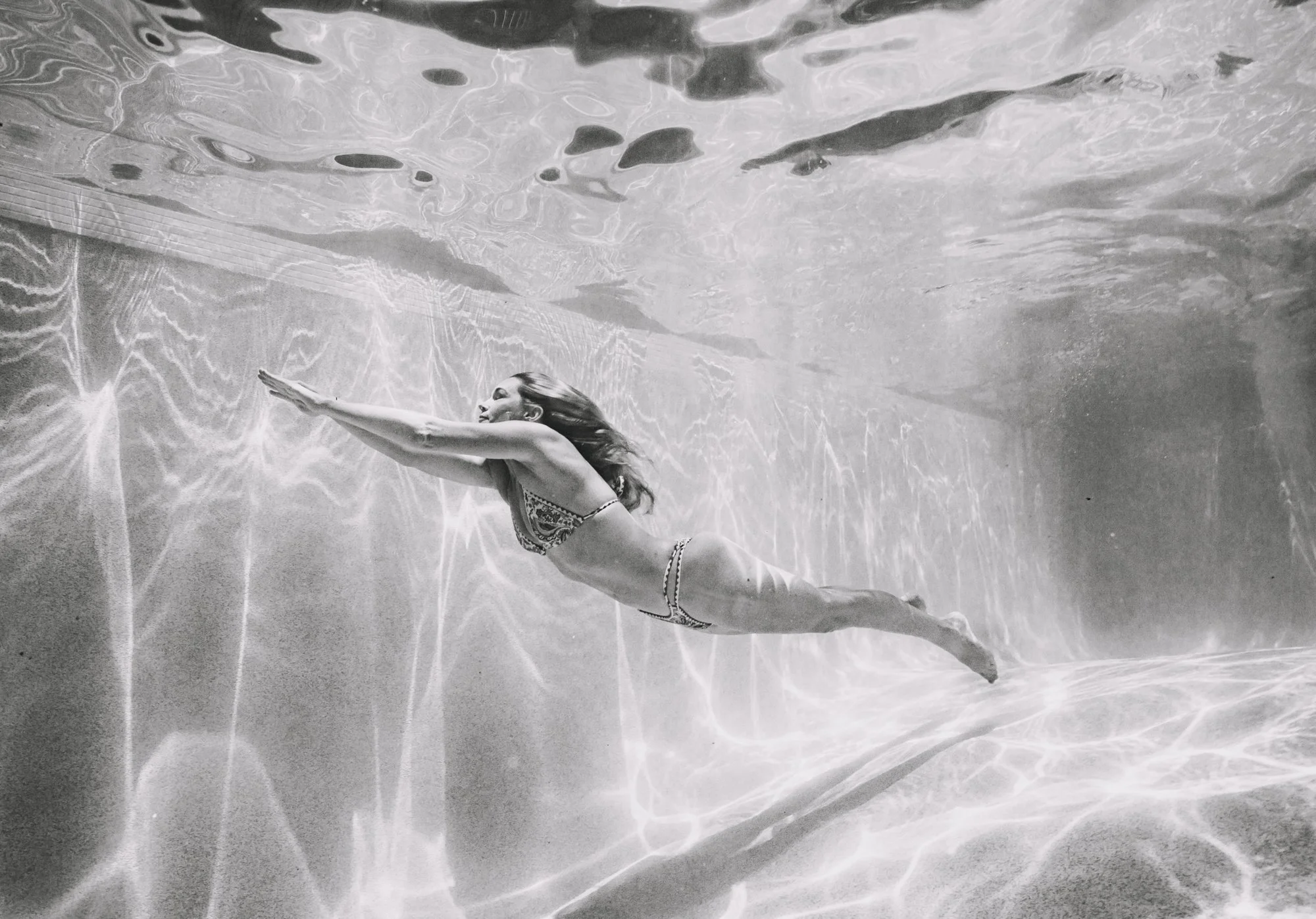 A woman in a bikini swimming underwater in a pool with her arms extended forward.