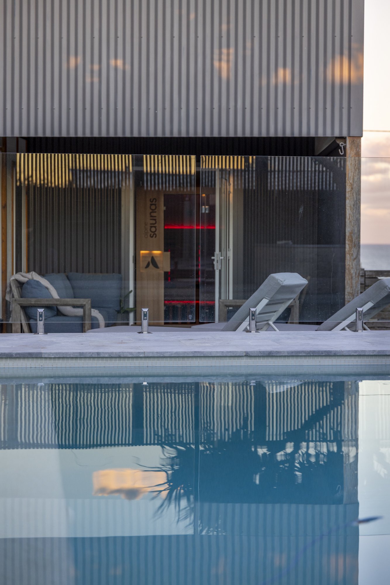 An outdoor swimming pool with clear blue water in front of a modern building with a glass wall, overlooking the ocean at sunset.