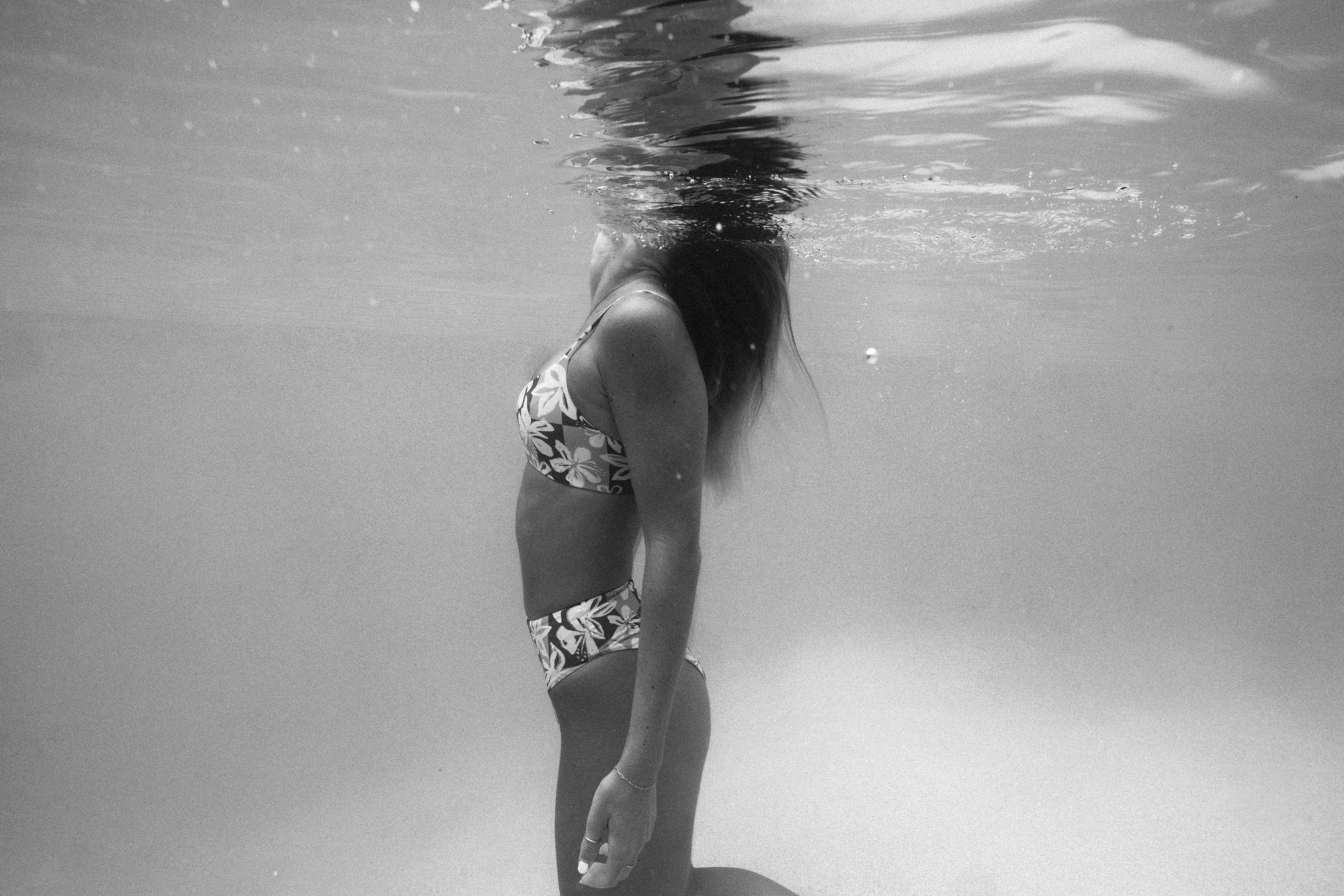 Black and white underwater photo of a woman wearing a floral bikini, with her head partly submerged and body visible underwater.