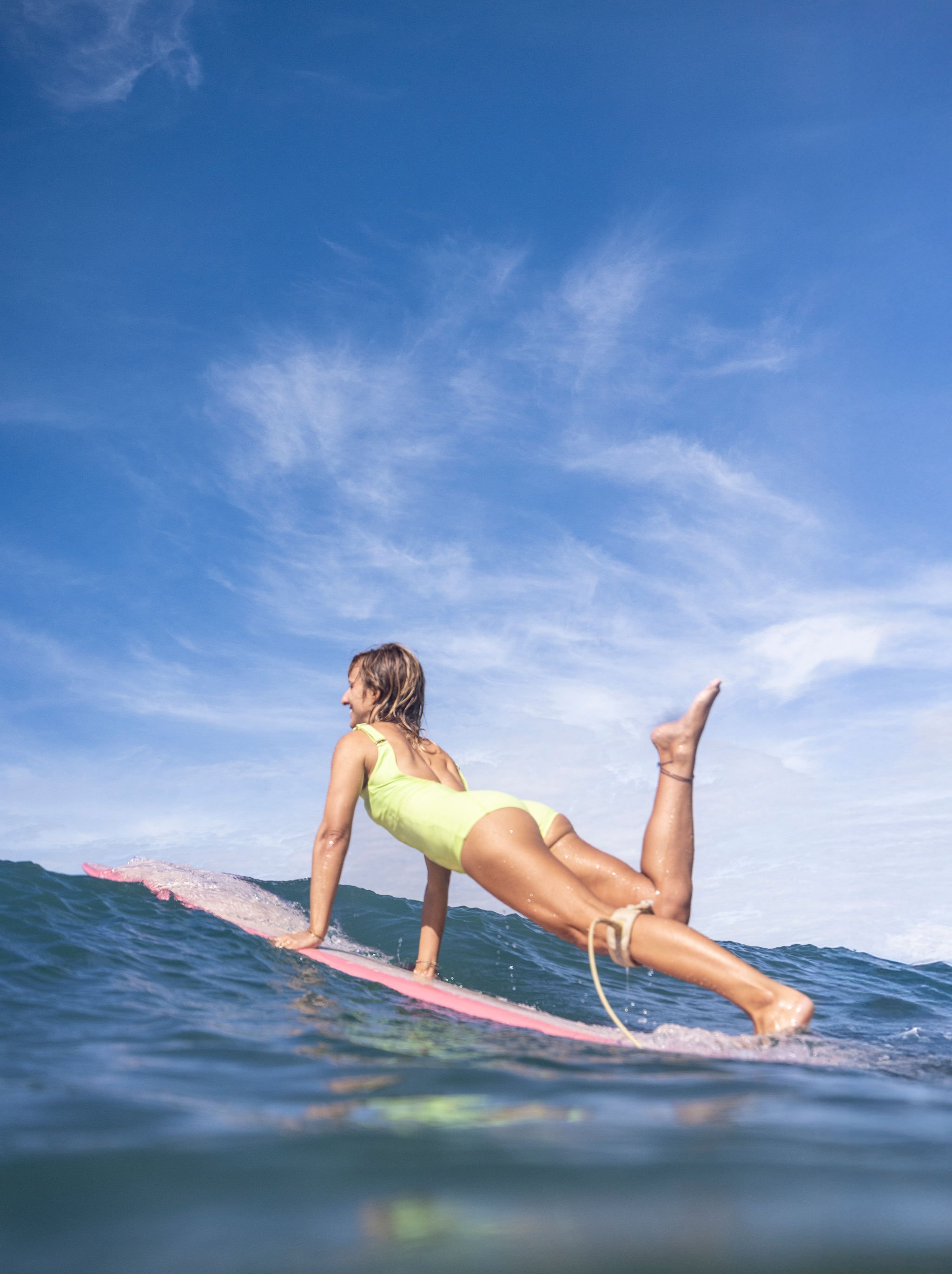 A woman in a yellow swimsuit riding a surfboard on the ocean with a blue sky and wispy clouds overhead.