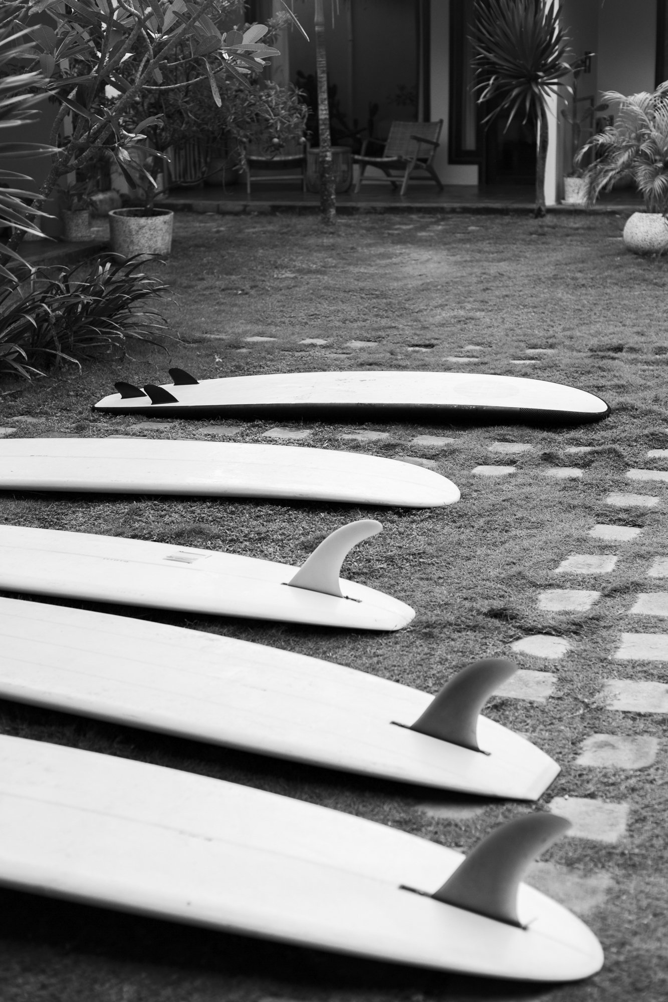Several surfboards with fins arranged on a grassy yard area near a patio with potted plants and outdoor furniture.