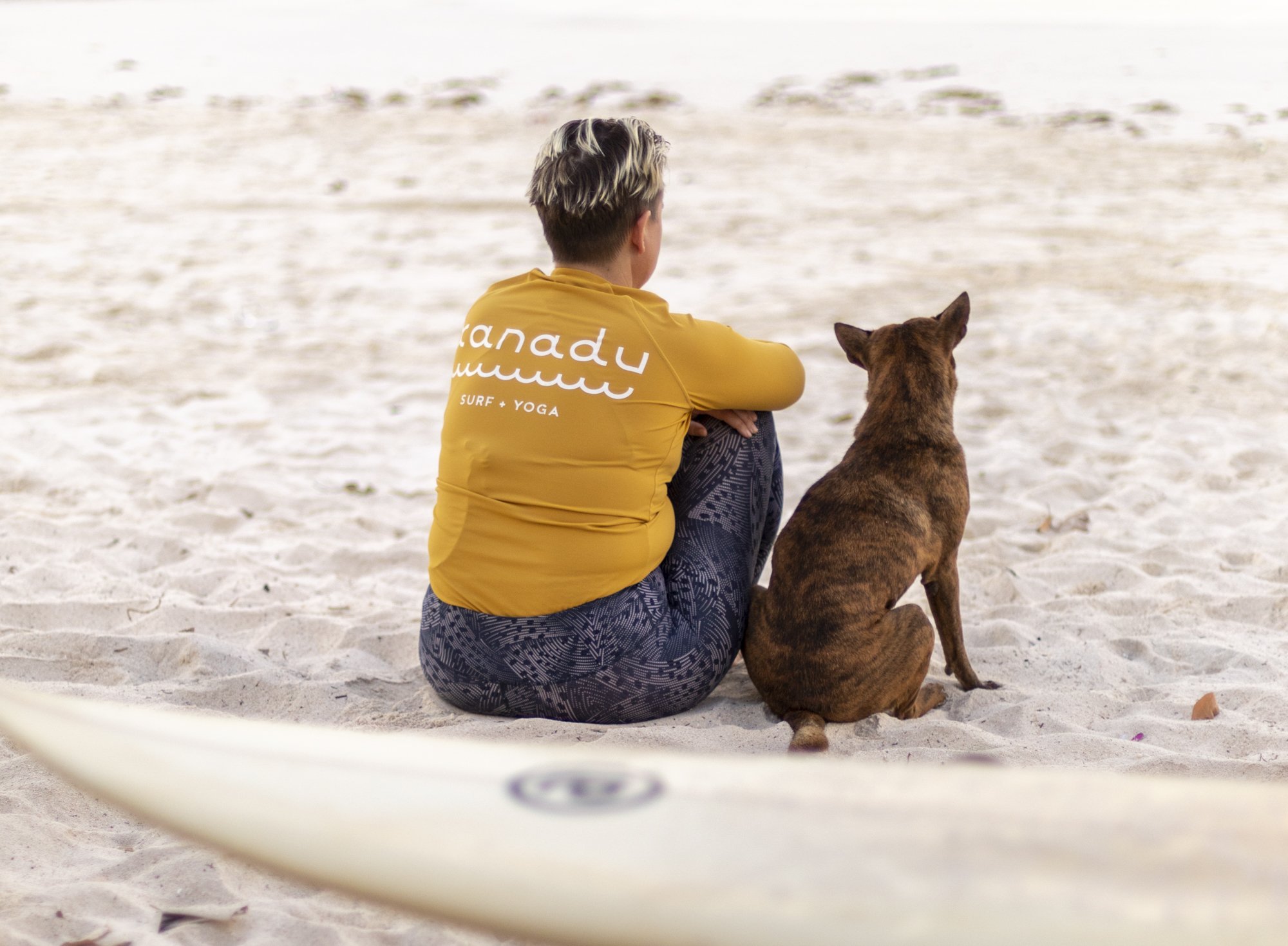 Person with short blond hair sitting on the sand with a brown dog, facing the ocean, on a beach.